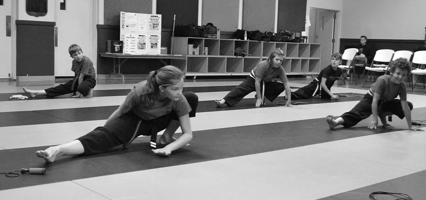 Group of children in a martial arts class stretching on mats in a gym.