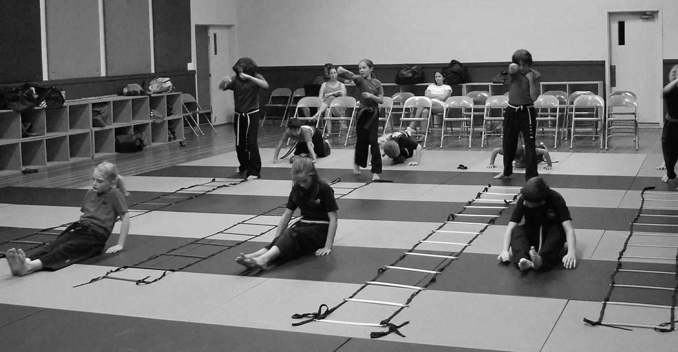 Children participating in a martial arts training session, practicing agility drills with ladders and hopscotch on mats inside a gymnasium.