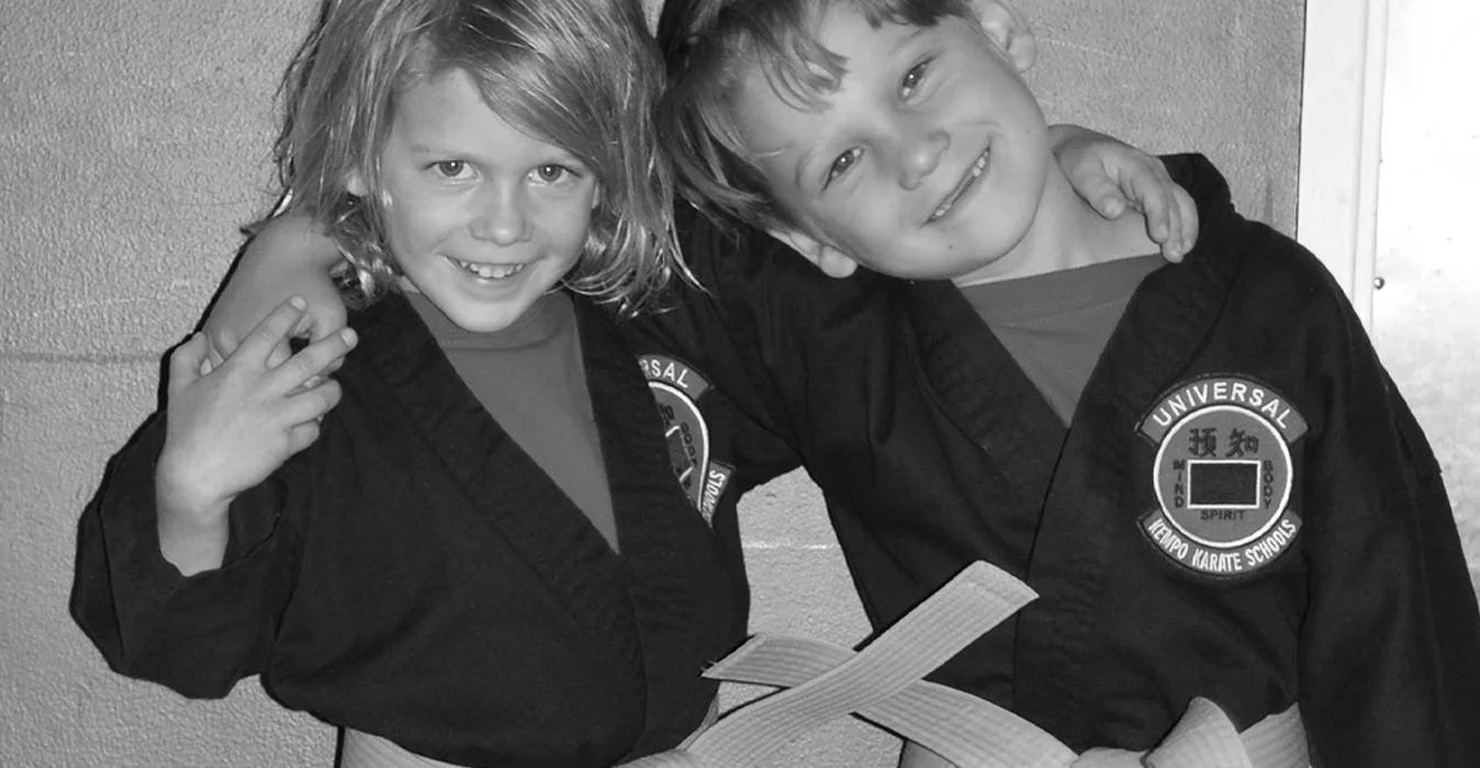 Two children in martial arts uniforms with patches, smiling and posing together, one with an arm around the other, against a textured wall.