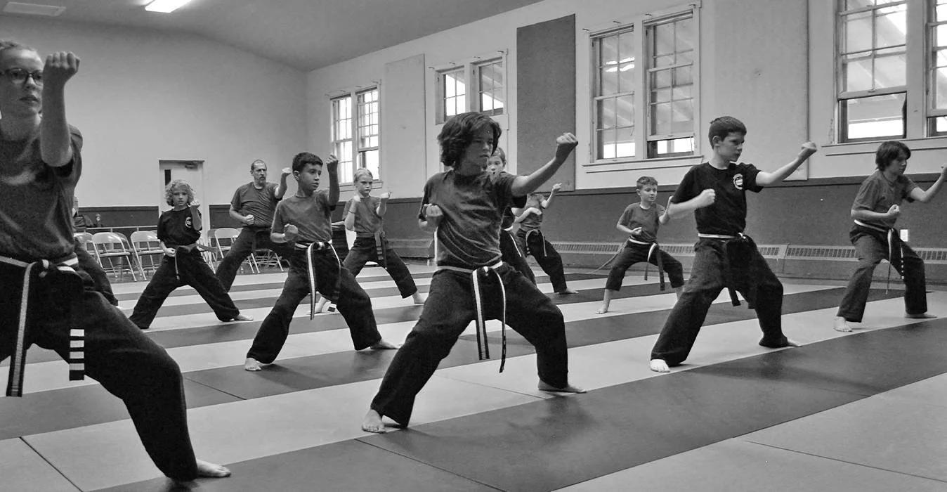 Kids in martial arts uniforms practicing punches in a classroom with padded floors and large windows.