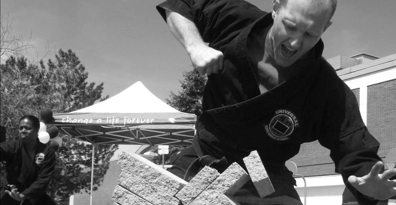 Professor Greenway in a martial arts uniform practicing breaking bricks during an outdoor event with a canopy tent and trees in the background.