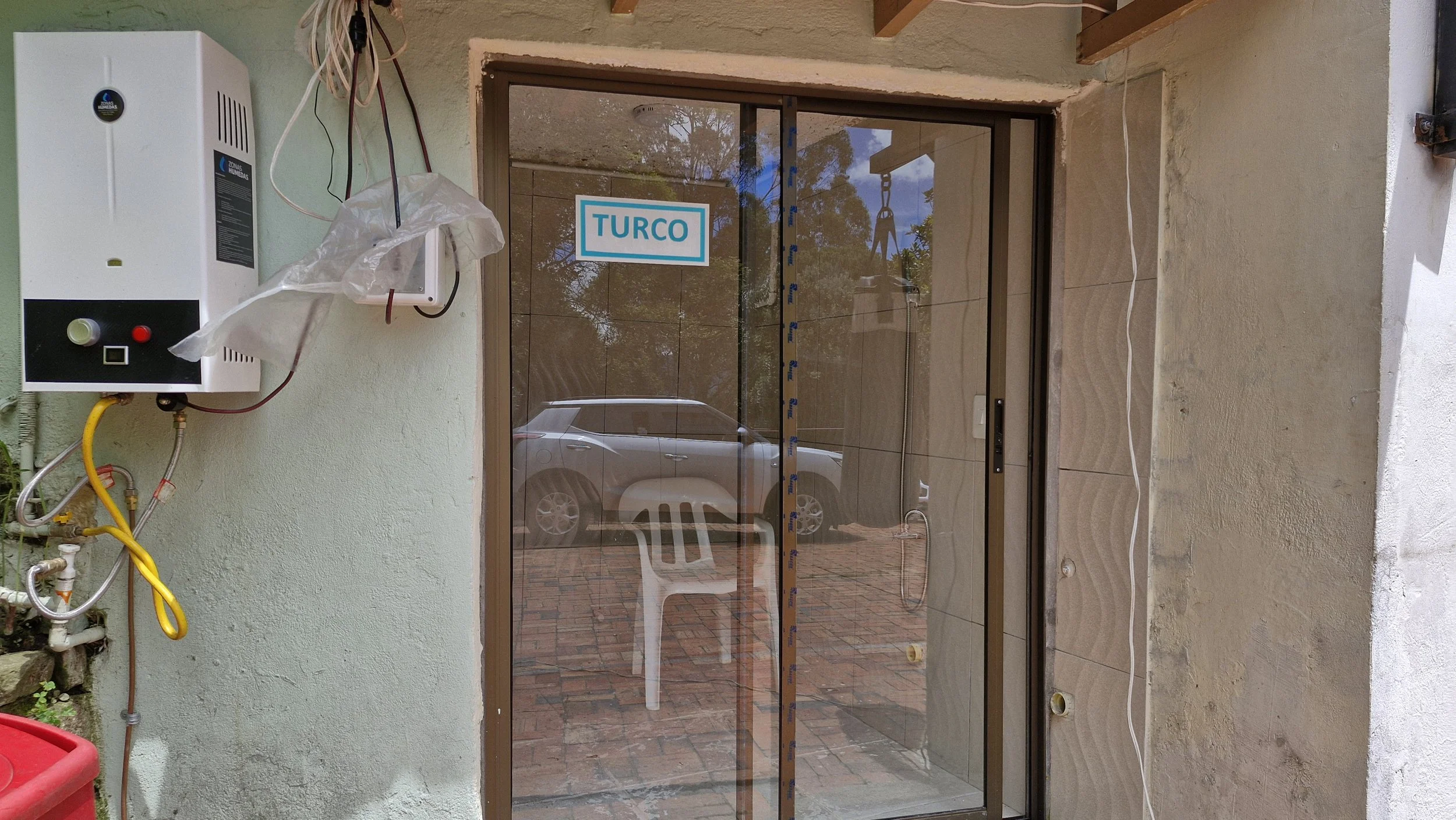 Puerta de vidrio con marco metálico y un letrero azul que dice 'TURCO'. Al lado hay un calentador de agua y algunos cables en la pared. Cerca de la puerta hay una silla plástica blanca y un coche reflejado en el vidrio.