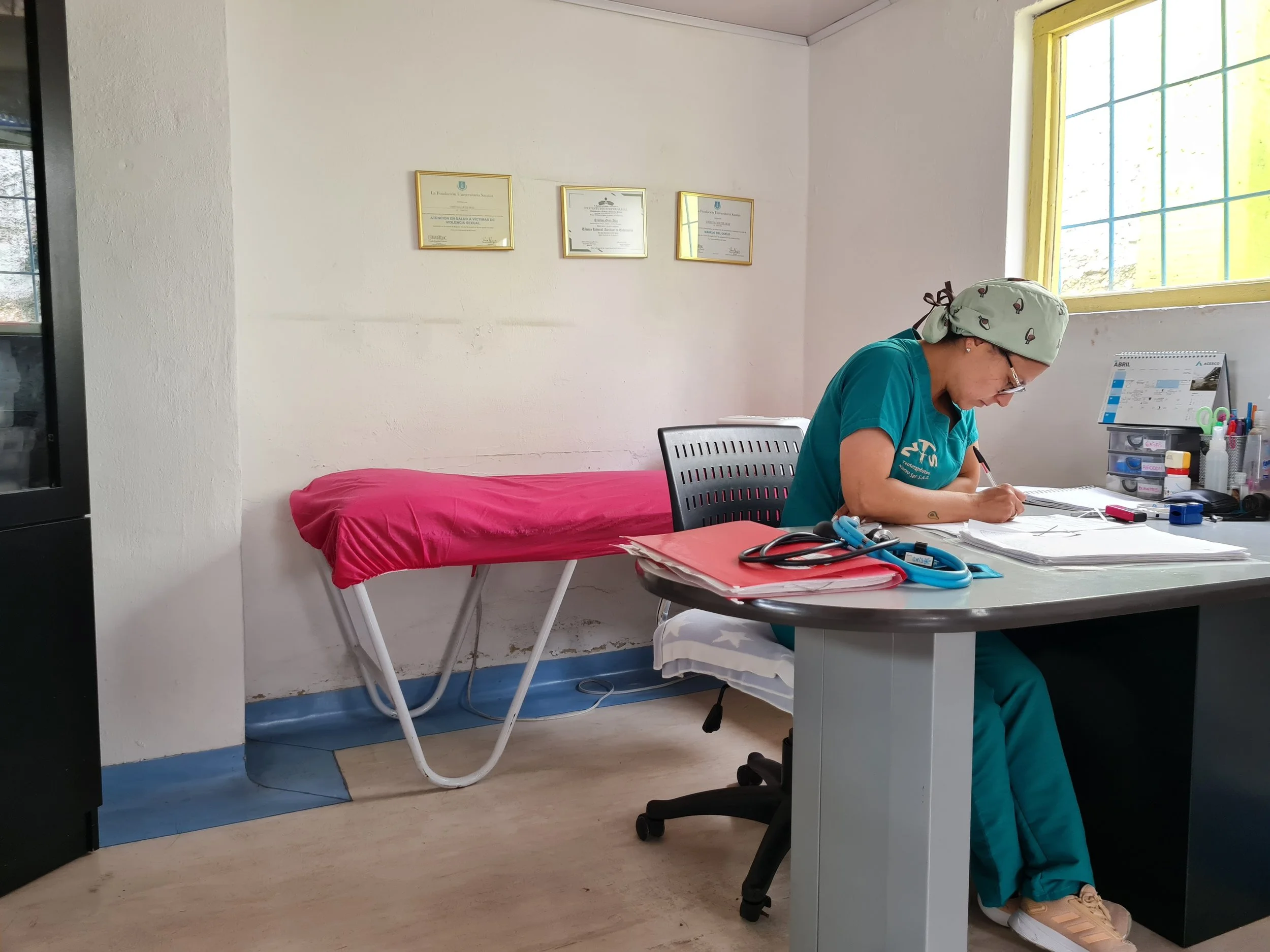 Una mujer con gafas, gorro y uniforme médico azul, sentada en una oficina, revisando documentos en una mesa de trabajo, con un escritorio que tiene instrumentos médicos y papeles. En la pared, hay tres diplomas enmarcados. Cerca de la ventana, hay una cama con sábana rosa.