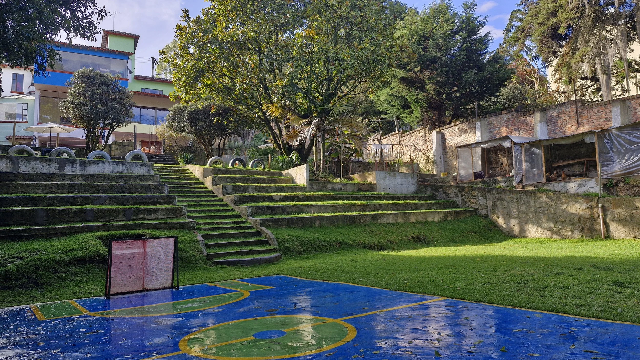 Área de juegos al aire libre con escaleras de piedra cubiertas de musgo, árboles grandes y un edificio colorido en el fondo.