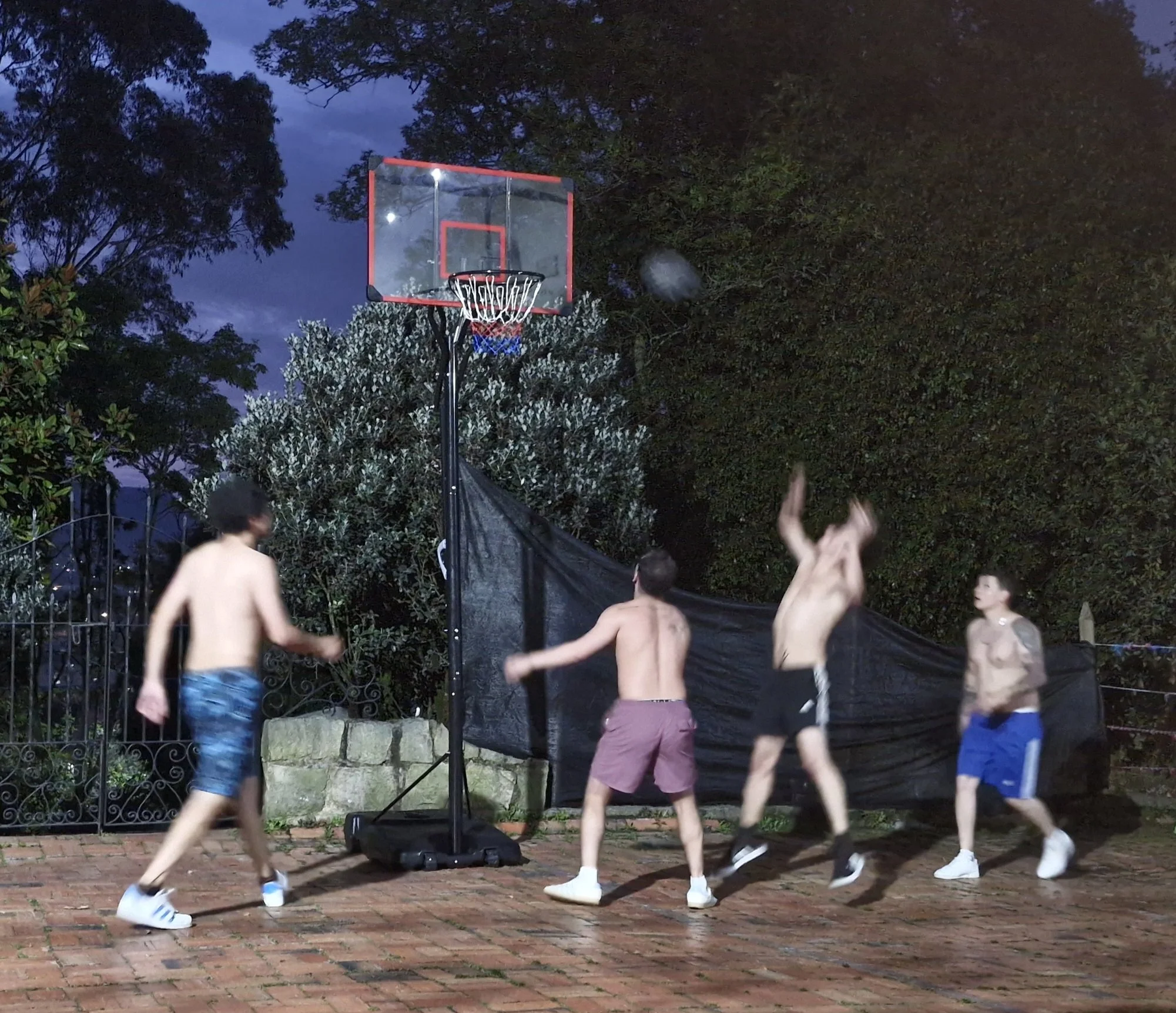 Niños jugando baloncesto sin ropa en un patio exterior por la noche, algunos corriendo, otros saltando y uno lanzando la pelota hacia la canasta.