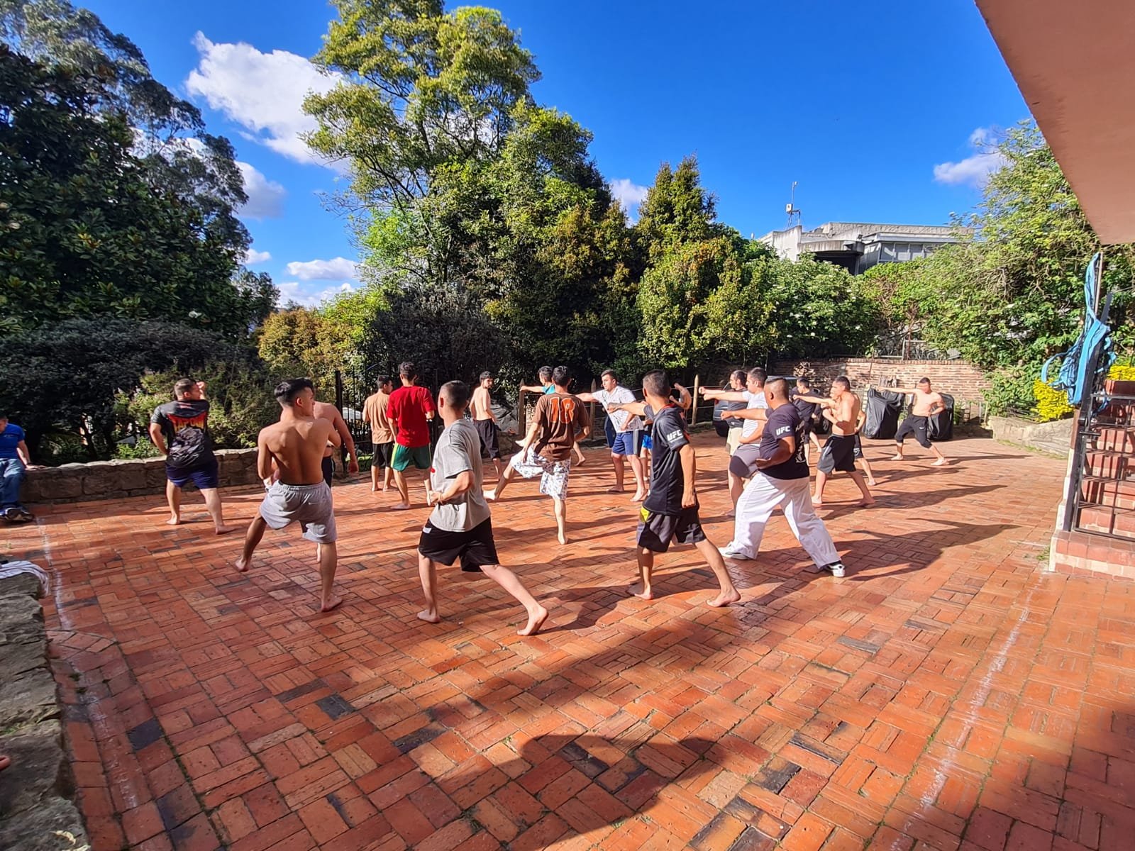 Grupo de personas practicando artes marciales en un patio de ladrillos exteriores con árboles verdes y un cielo azul.