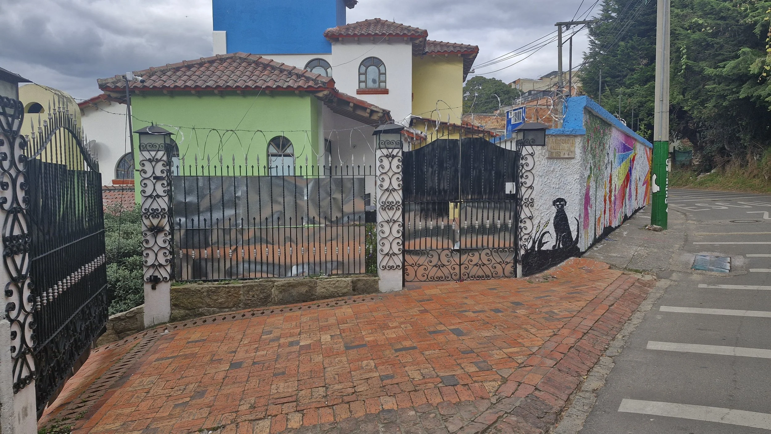 Casa pintada con mural colorido en la pared con un perro negro en la esquina, rejas negras y un camino de ladrillos.