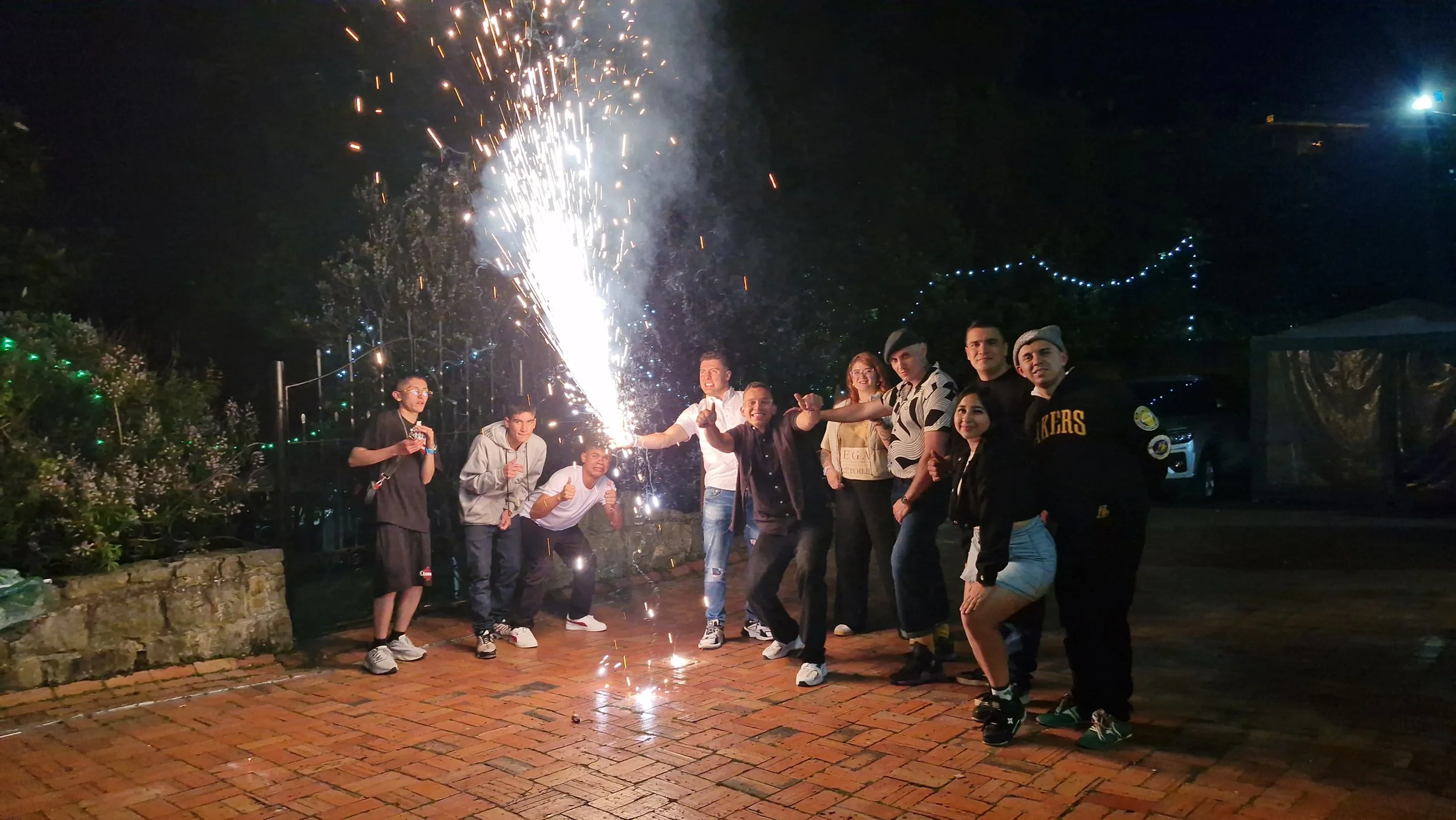 Un grupo de personas celebrando en la noche con fuegos artificiales, sonriendo y posando para la foto en un espacio al aire libre decorado con luces.