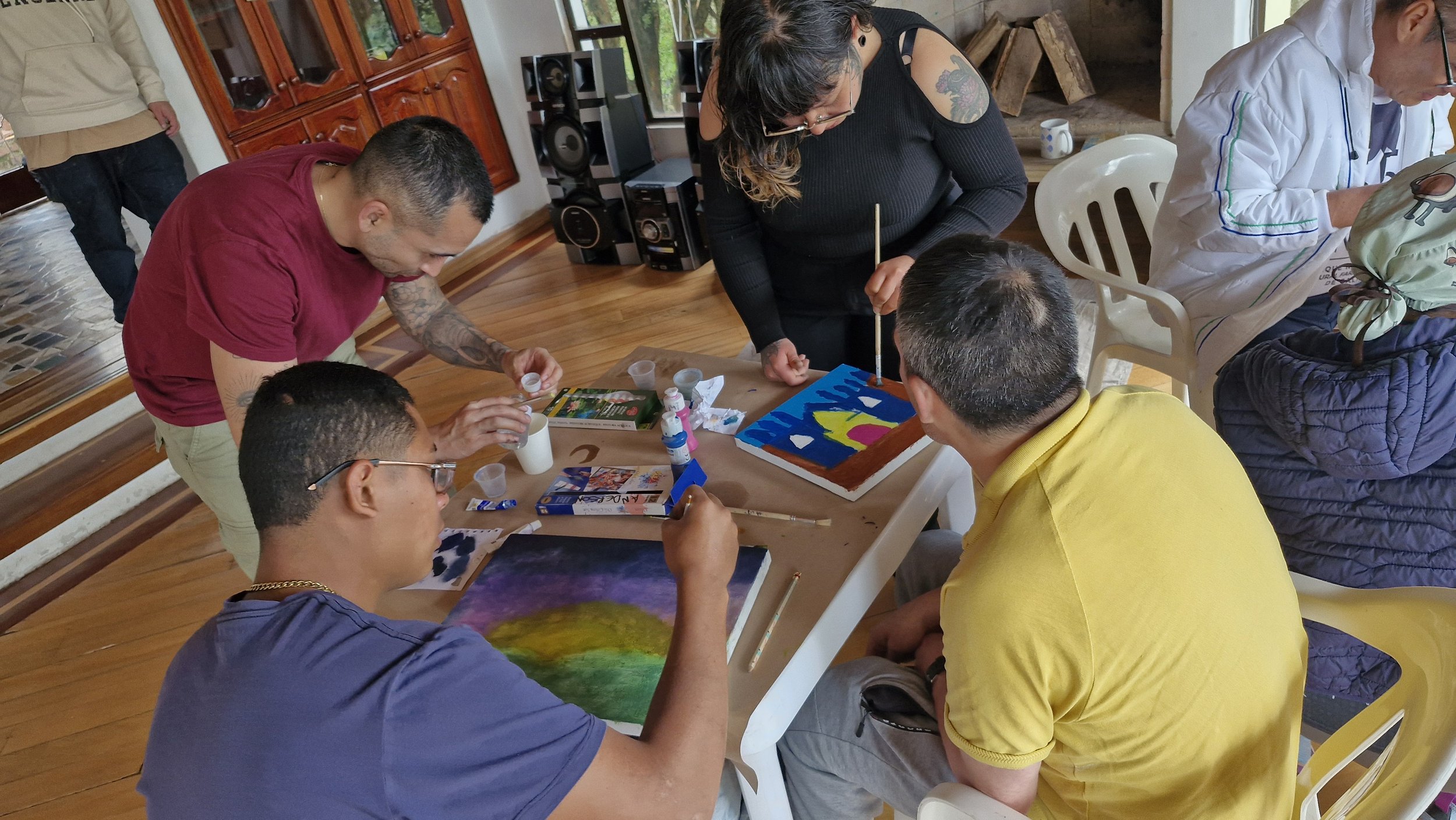 Varias personas trabajando en un taller de pintura en una mesa con materiales de arte, en un ambiente interior con madera y ventanas grandes.