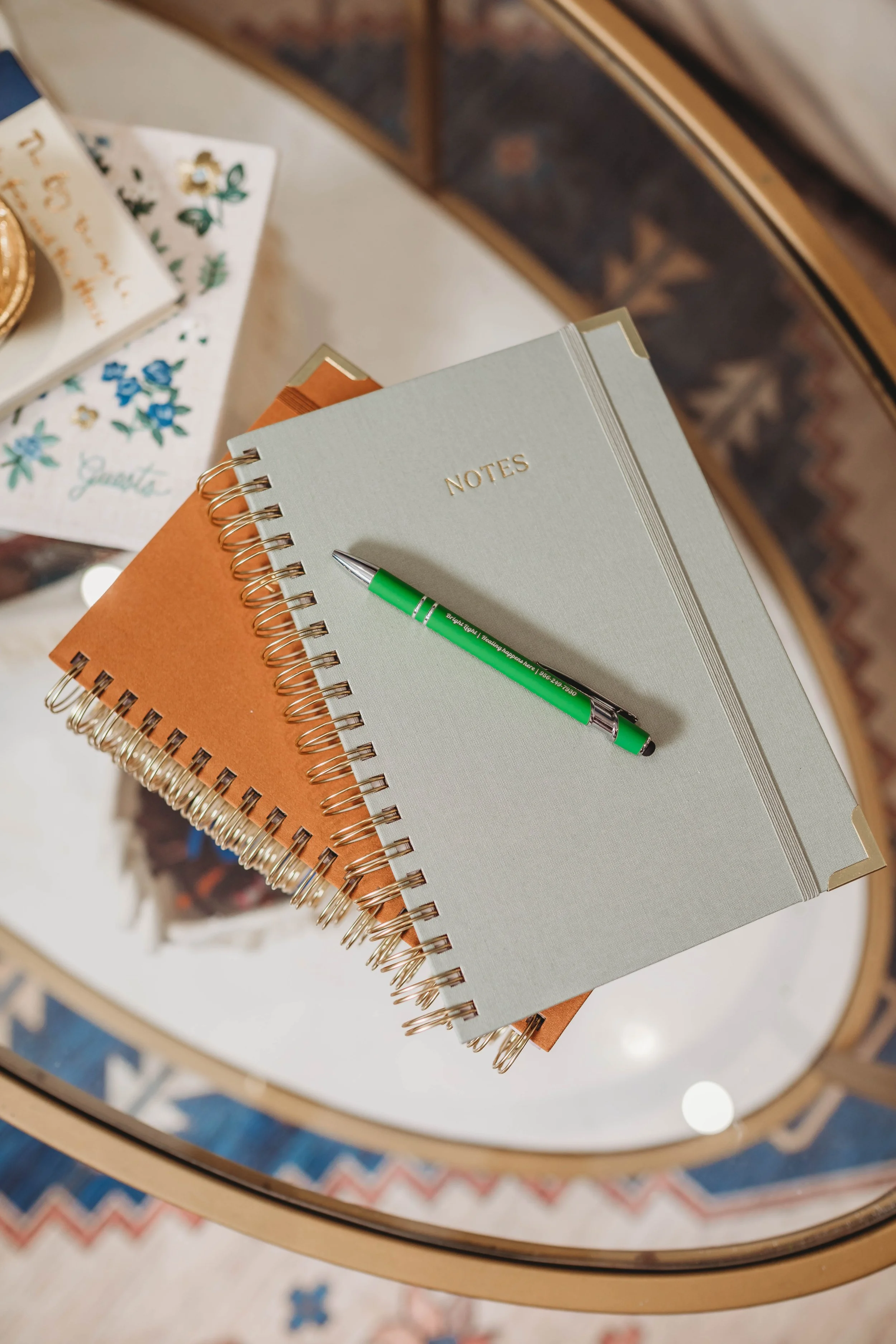 A close-up view of a desk with three spiral-bound notebooks stacked on top of each other; the top notebook is light green with the word 'NOTES' embossed in gold; a green and silver pen resting on it; in the background, partially visible, is a white book with floral designs.
