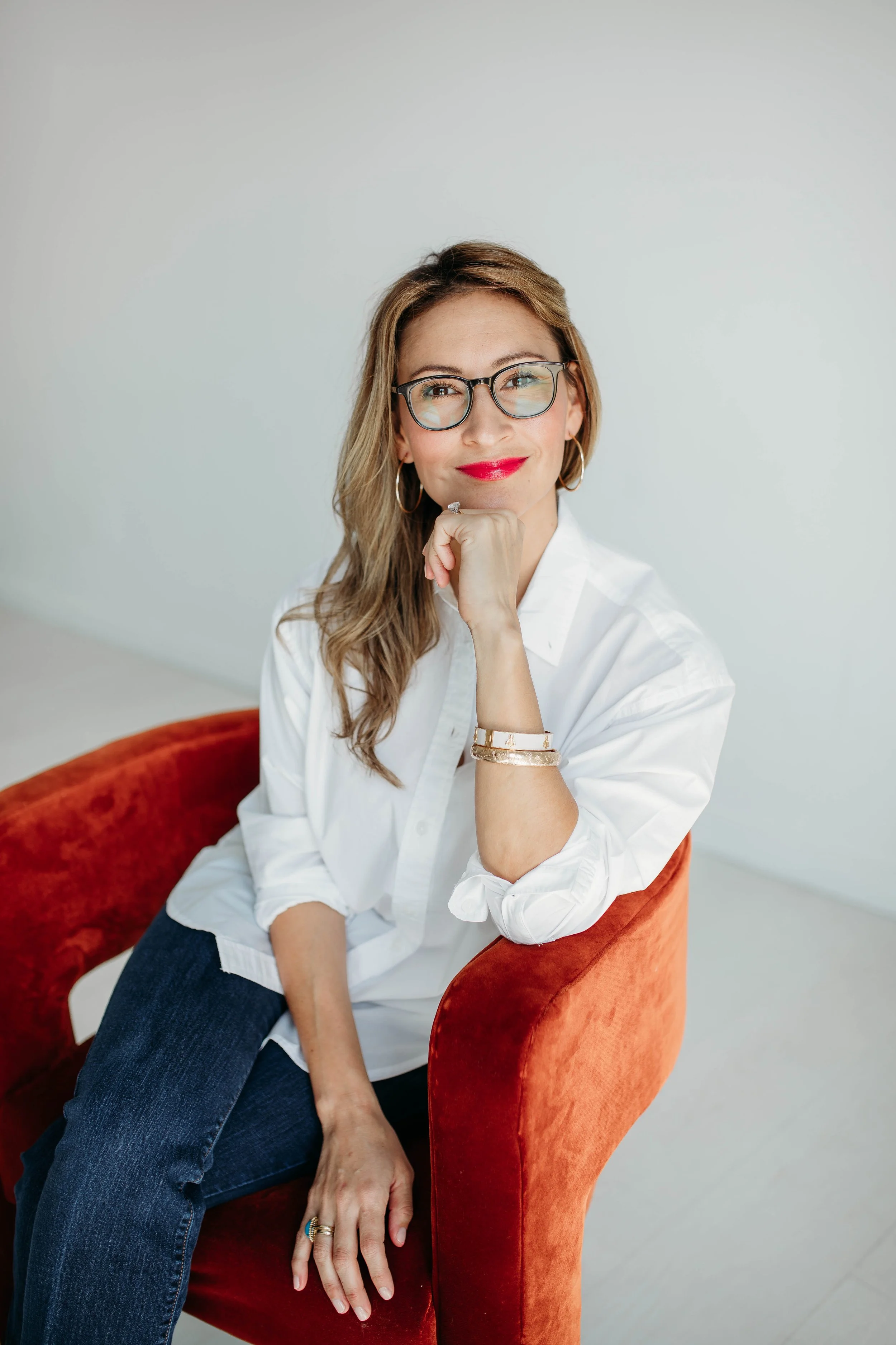 A woman with glasses and red lipstick sitting on an orange velvet chair, resting her chin on her hand, dressed in a white shirt and dark jeans, against a plain light grey background.