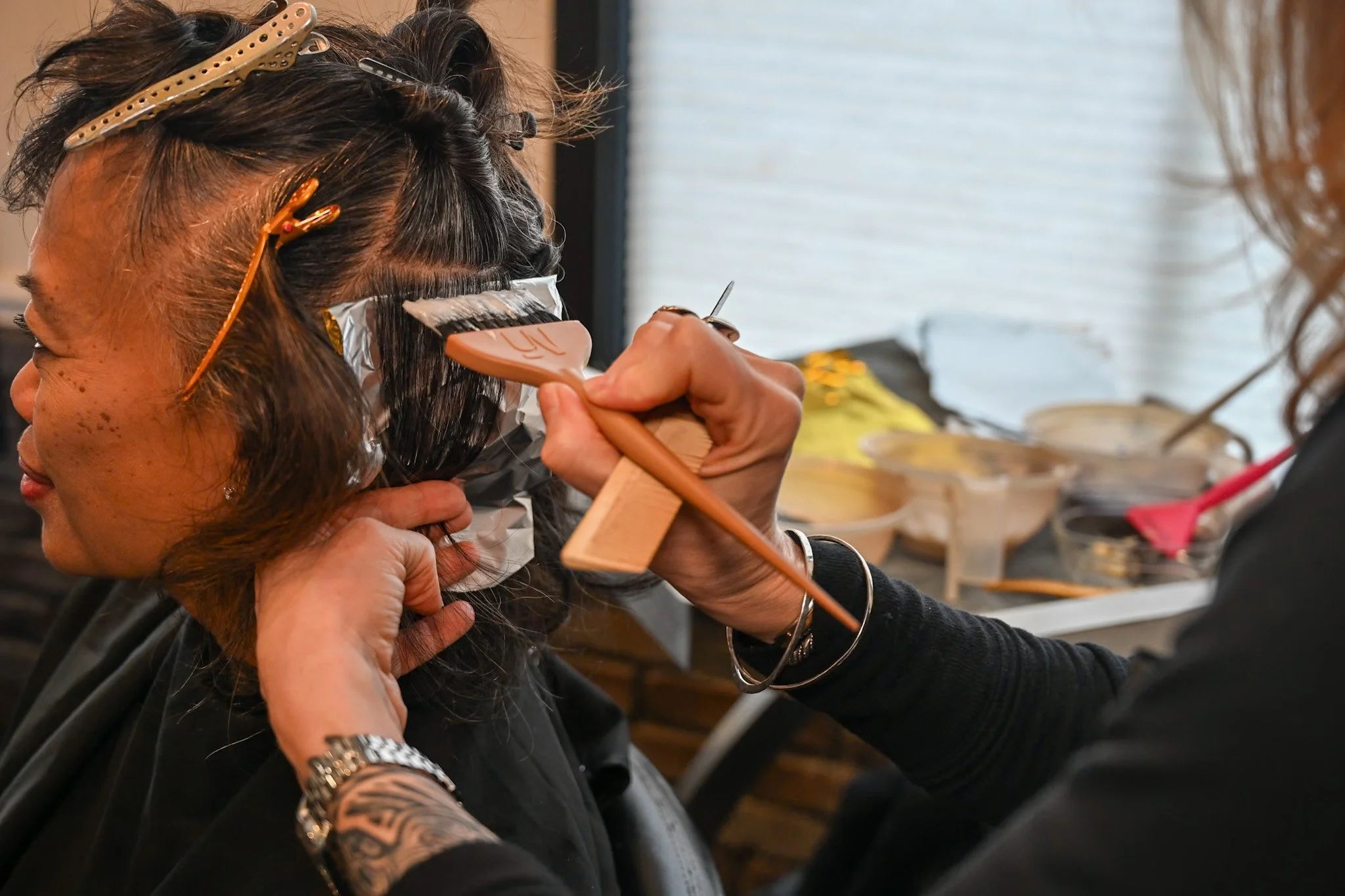 A woman getting her hair dyed by a stylist in a salon. The stylist is applying dye with a brush and wearing black clothes. The woman has dark hair, some of which is already covered in aluminum foil. The background shows various hair products and tools.