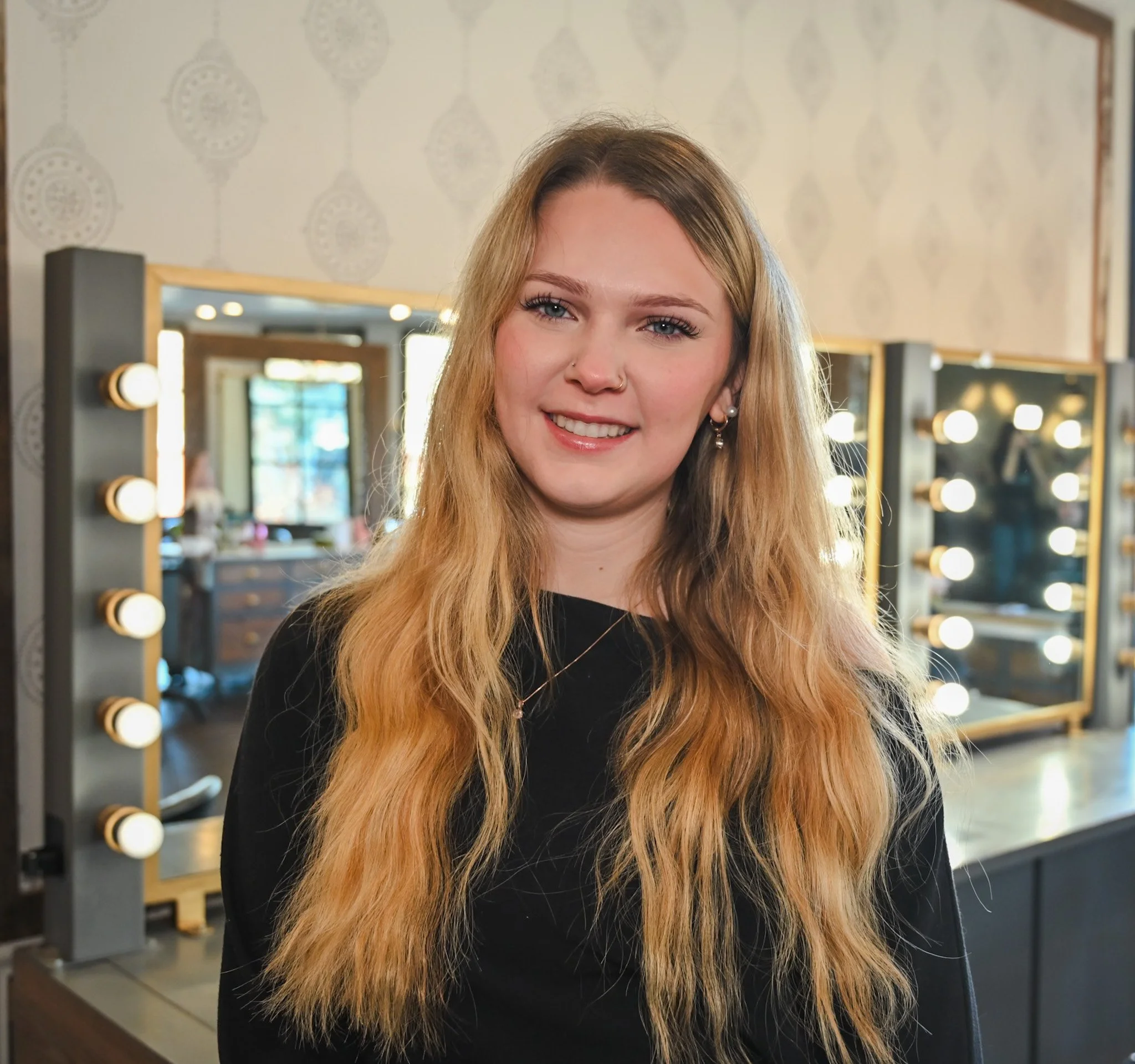 A smiling woman with long blonde hair, wearing earrings and a black top, standing in a room with large mirrors and light bulbs around the mirrors.