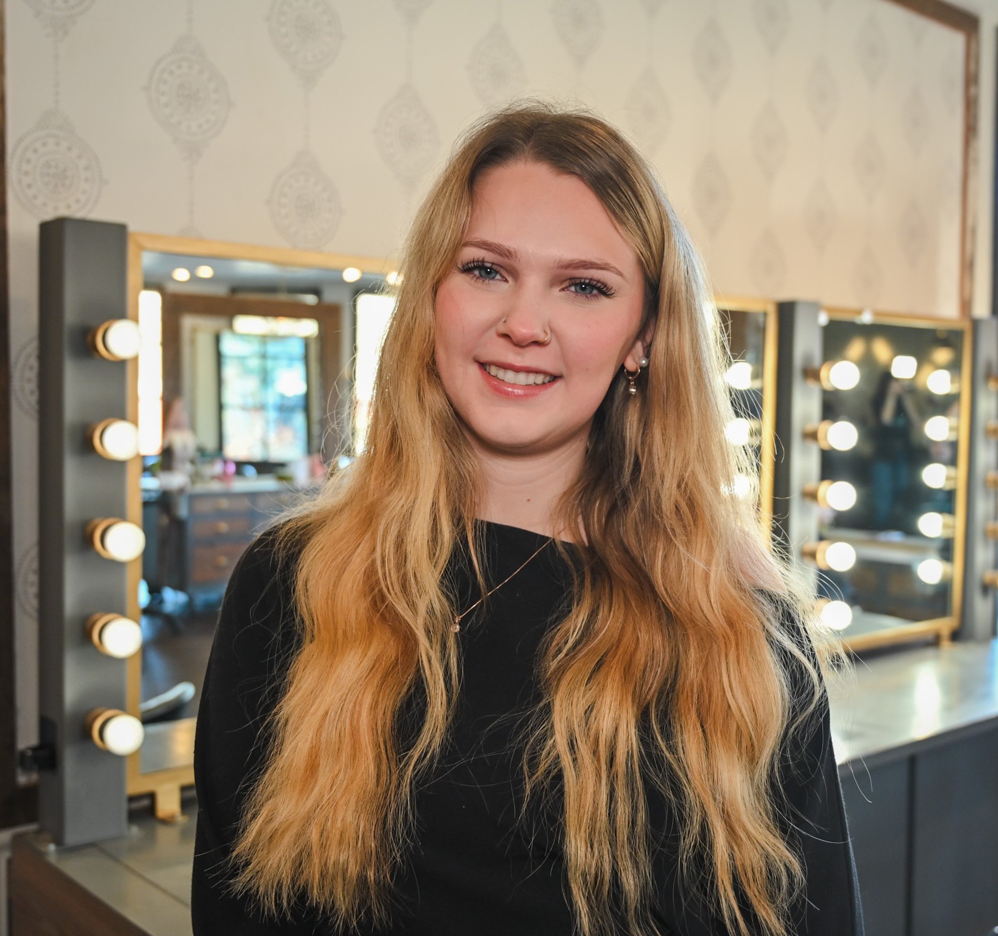 A young woman with long blonde hair and light skin smiling at the camera in a salon. She has a nose piercing and earrings. Behind her are mirrors and vanity lights.