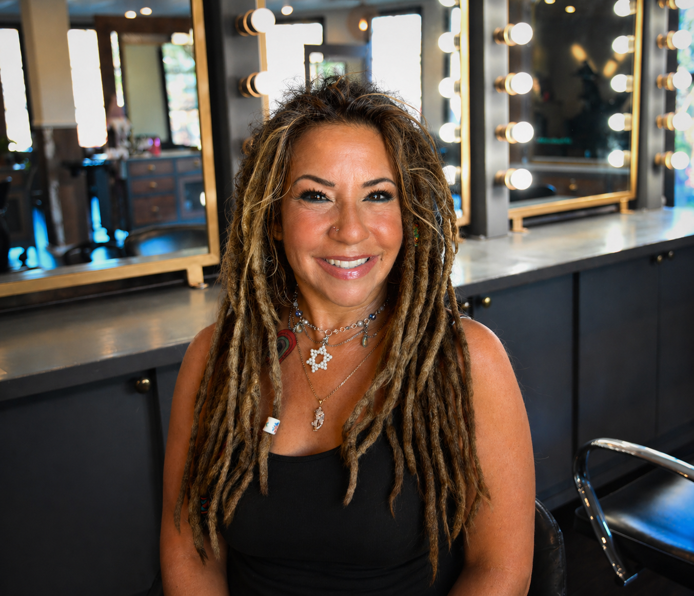 A woman with tan skin, long blonde dreadlocks, and colorful jewelry sitting in a well-lit salon or studio with mirrors and vanity lights in the background.