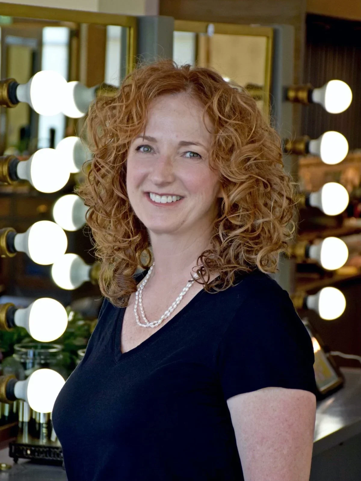 A smiling woman with curly red hair wearing a black top and pearl necklace, standing in front of a mirror with bright round lights.
