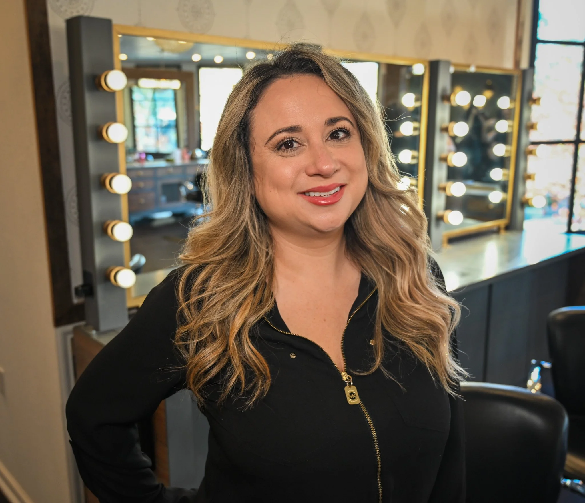 A woman with long, wavy blonde hair and wearing a black top with gold zipper, smiling in front of a makeup mirror with lights in a room that has large windows.