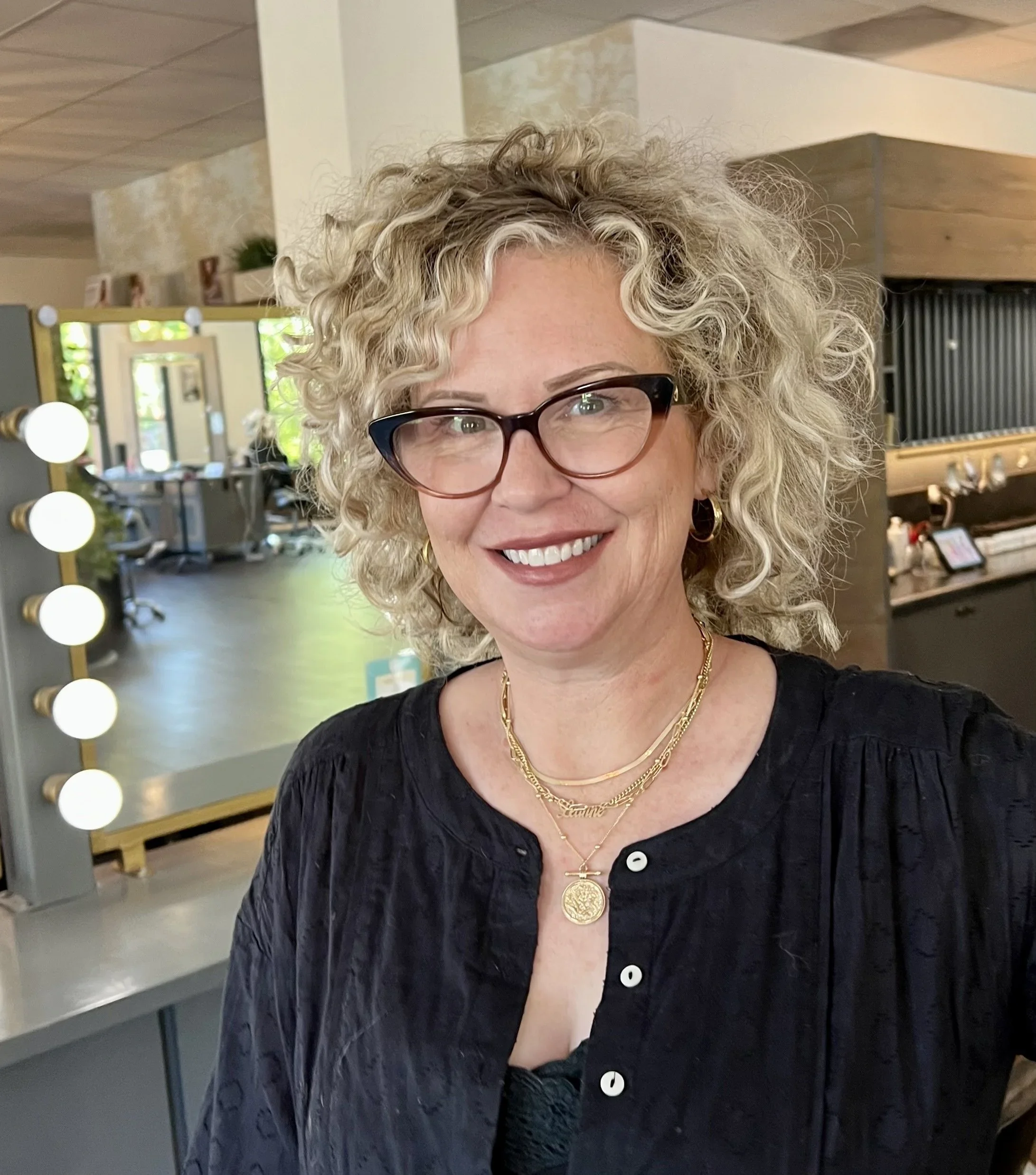 A woman with curly blonde hair, glasses, and gold jewelry smiling in a salon or studio.