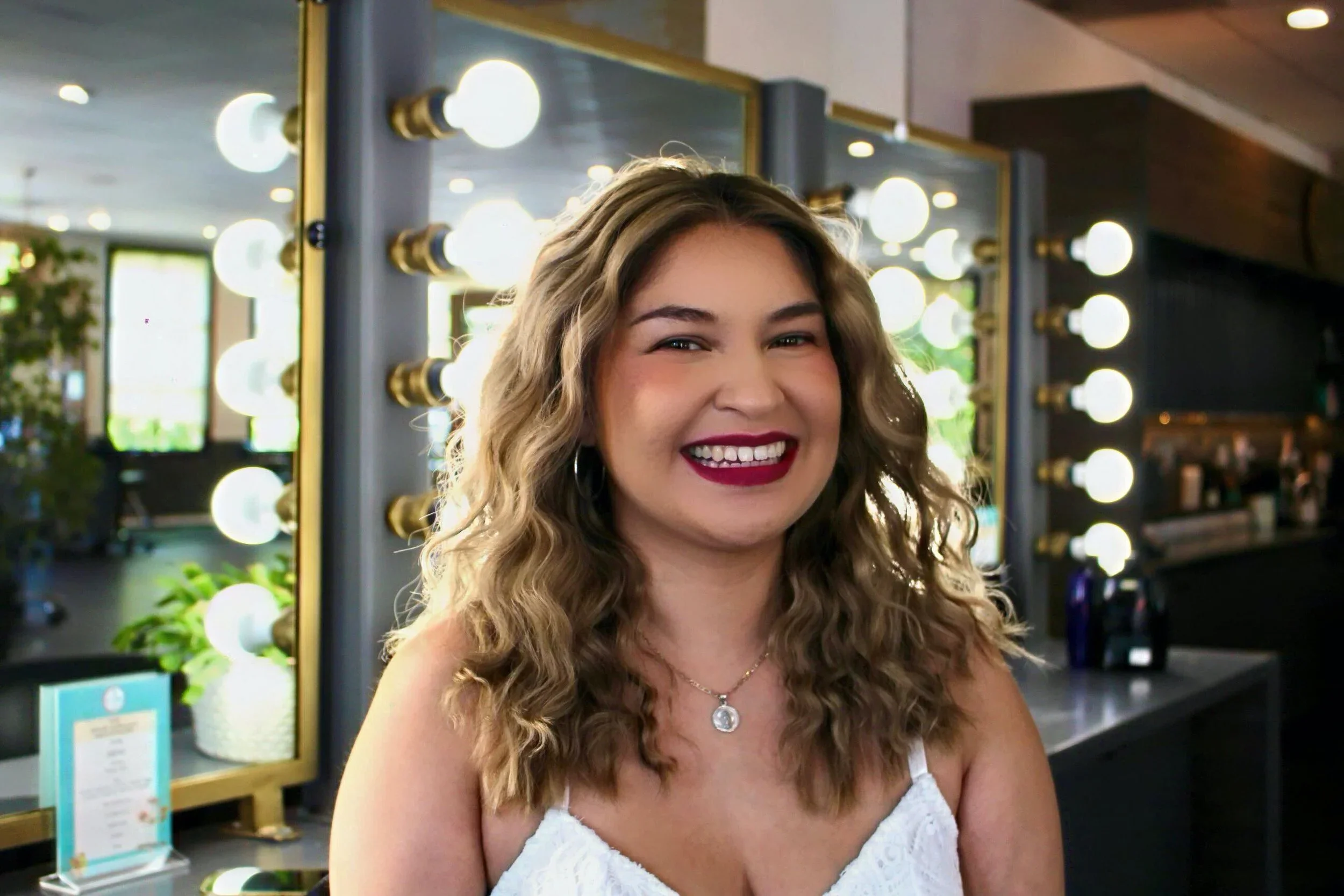 A woman with wavy blonde hair, wearing a white top and a necklace, smiling in front of a dressing mirror with bright lights inside a salon or makeup studio.