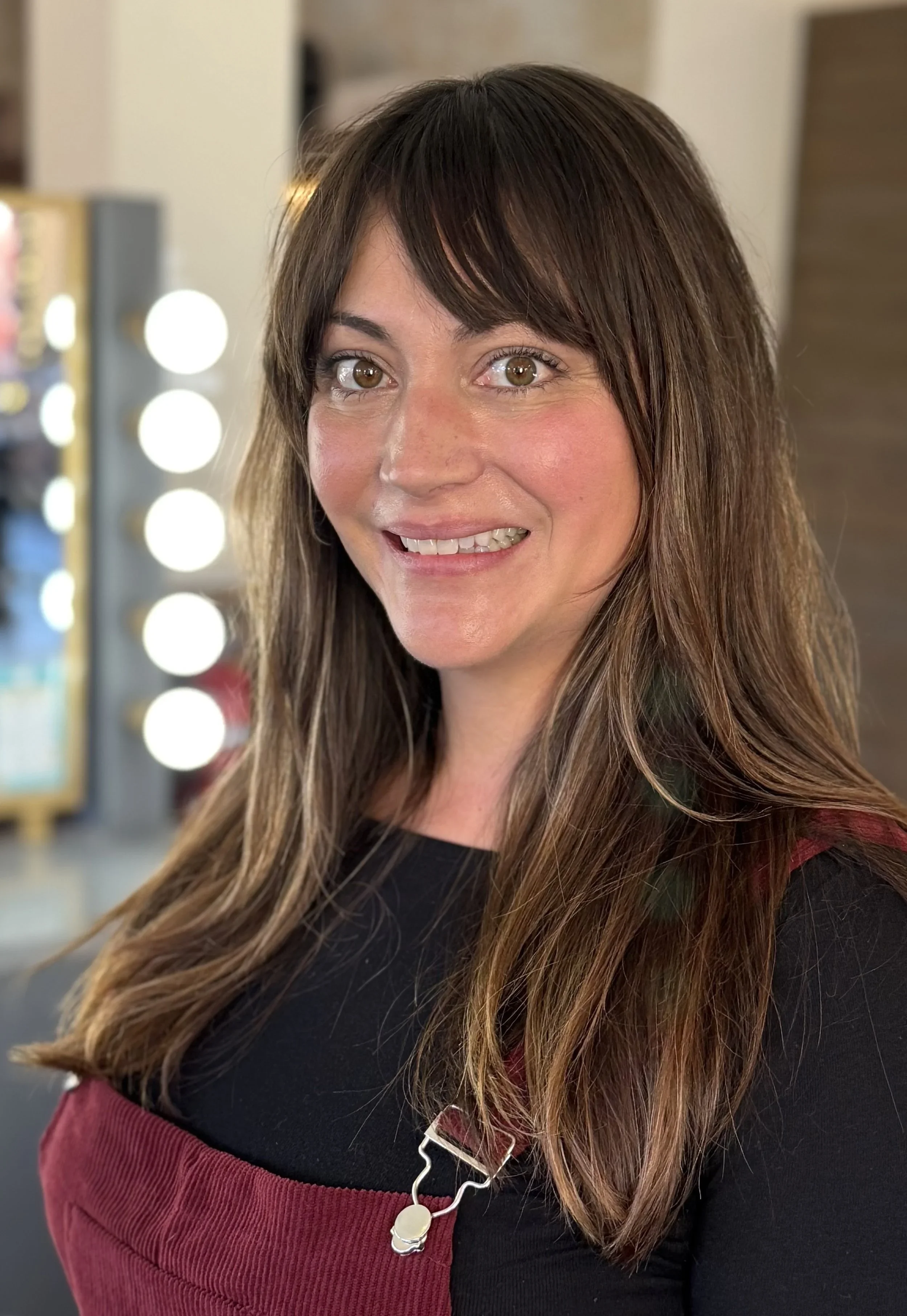 A woman with long brown hair and brown eyes smiling, wearing a black top and a maroon apron, standing indoors with a background of lights and a blurred setting.