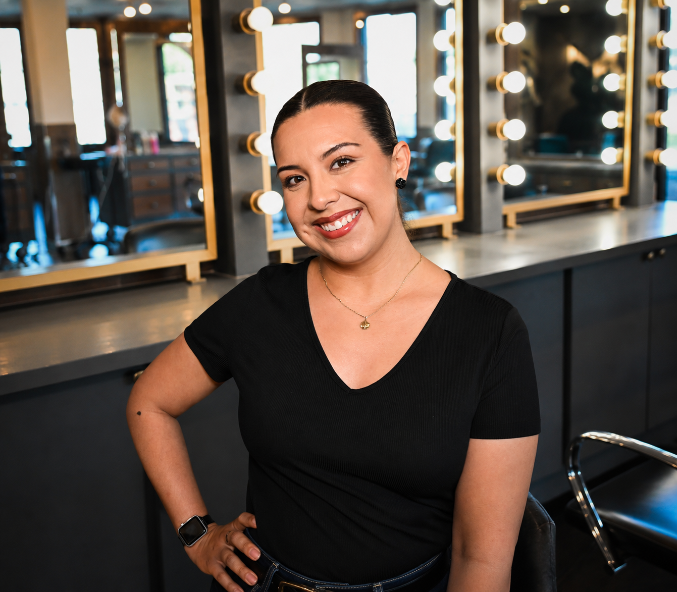 A woman with dark hair pulled back, wearing a black v-neck shirt, jewelry, and a smartwatch, standing in front of a mirror with vanity lights in a hair salon.