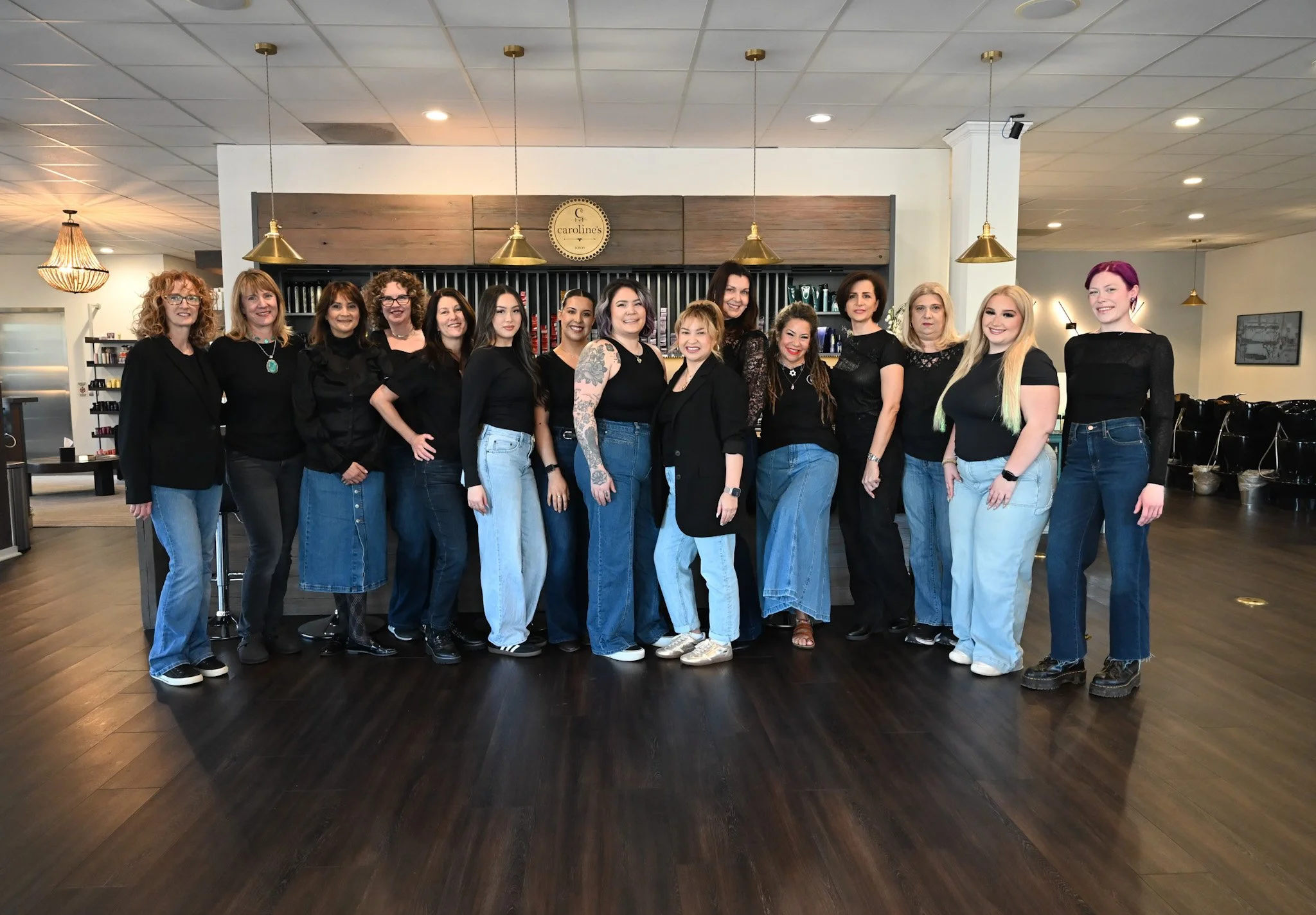 Group of 15 women standing together inside a hair salon, smiling at the camera, dressed in black tops and blue jeans.