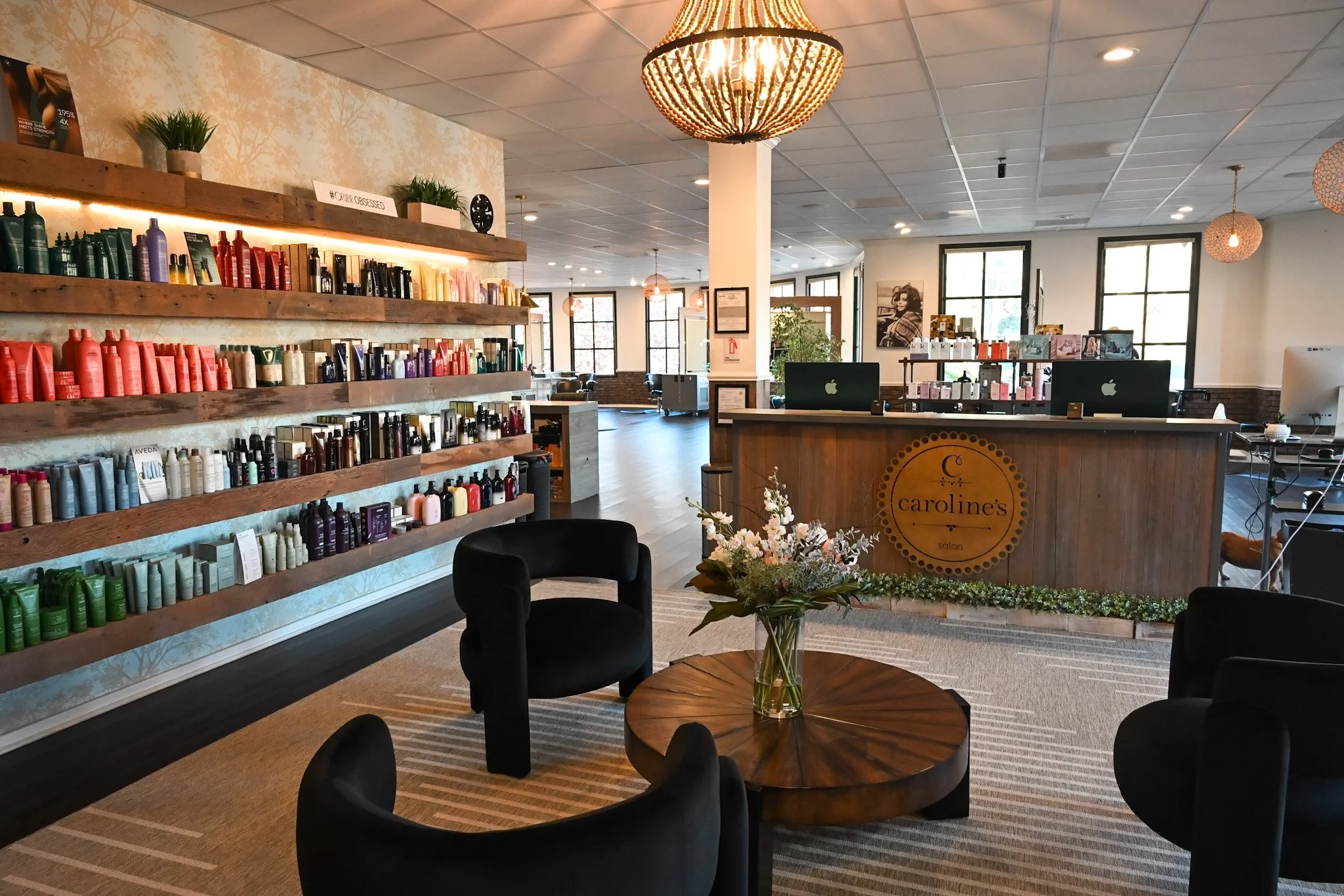Interior of a salon with a reception desk, black armchairs around a wooden table with a flower arrangement, and shelves of colorful hair care products on the wall.