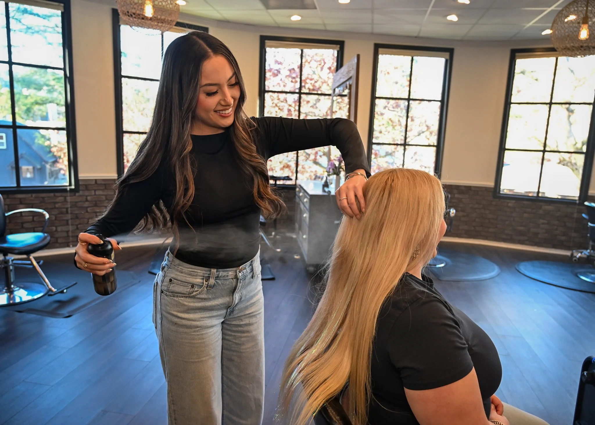 A woman with long dark hair styling a blonde woman's hair in a salon with large windows, modern decor, and black chairs.