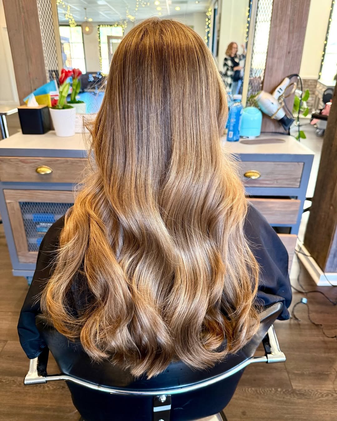 A woman with long, wavy, light brown hair sitting in a salon chair, with her back facing the camera. The salon has a mirror, plants, and styling tools visible in the background.