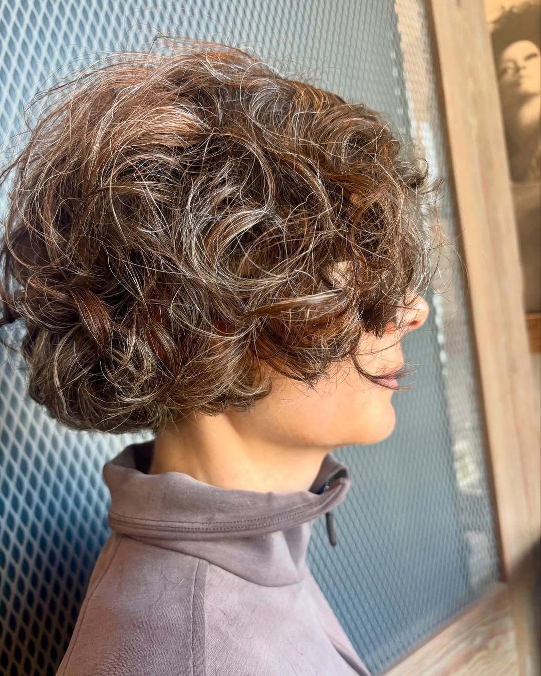 A woman with short, curly brown hair with highlights, wearing a gray turtleneck, standing outdoors near a wooden structure and metal fence.