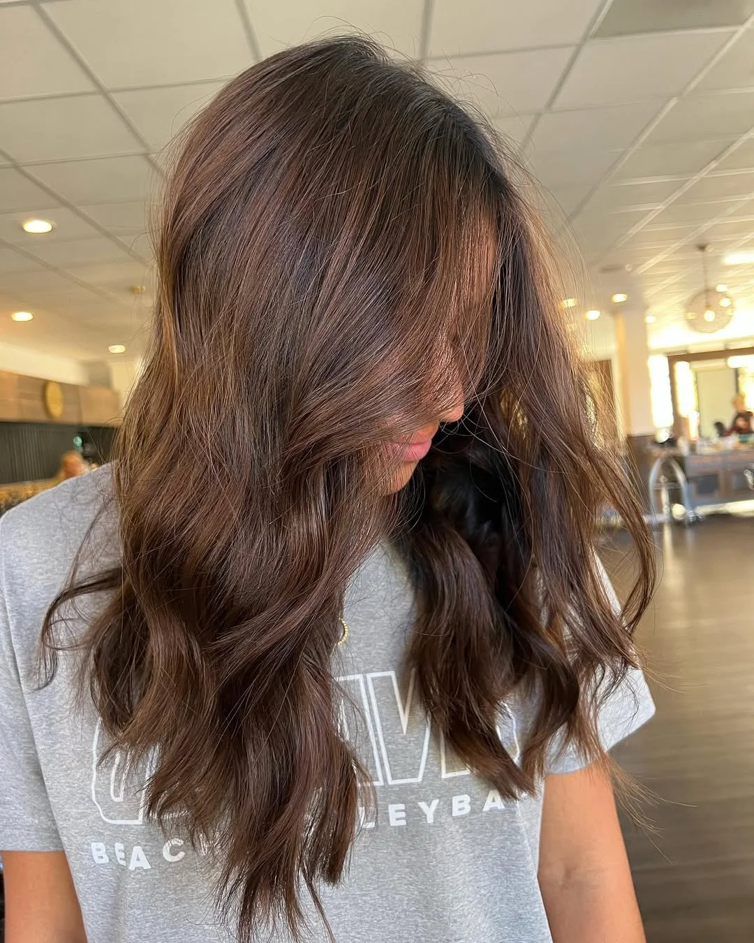 Woman with long wavy brown hair, looking down in a cafe or restaurant setting.