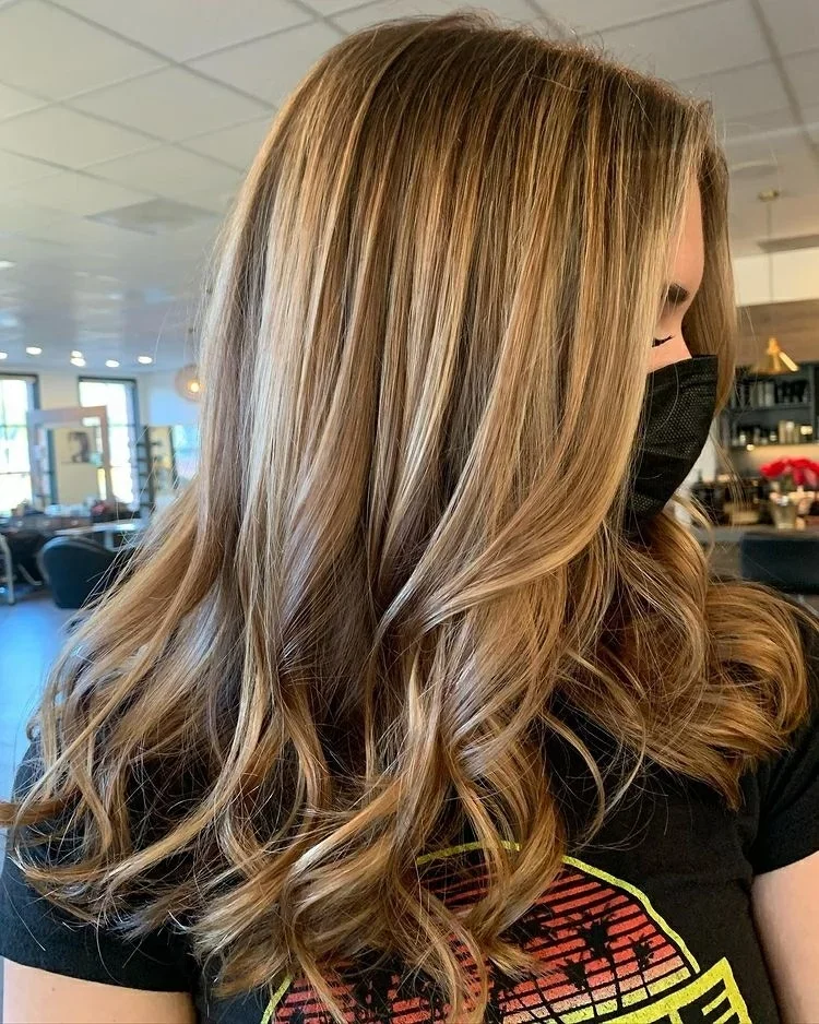 Side profile of woman with long, wavy blonde hair wearing a black face mask and a black t-shirt with a colorful graphic print inside a brightly lit room.