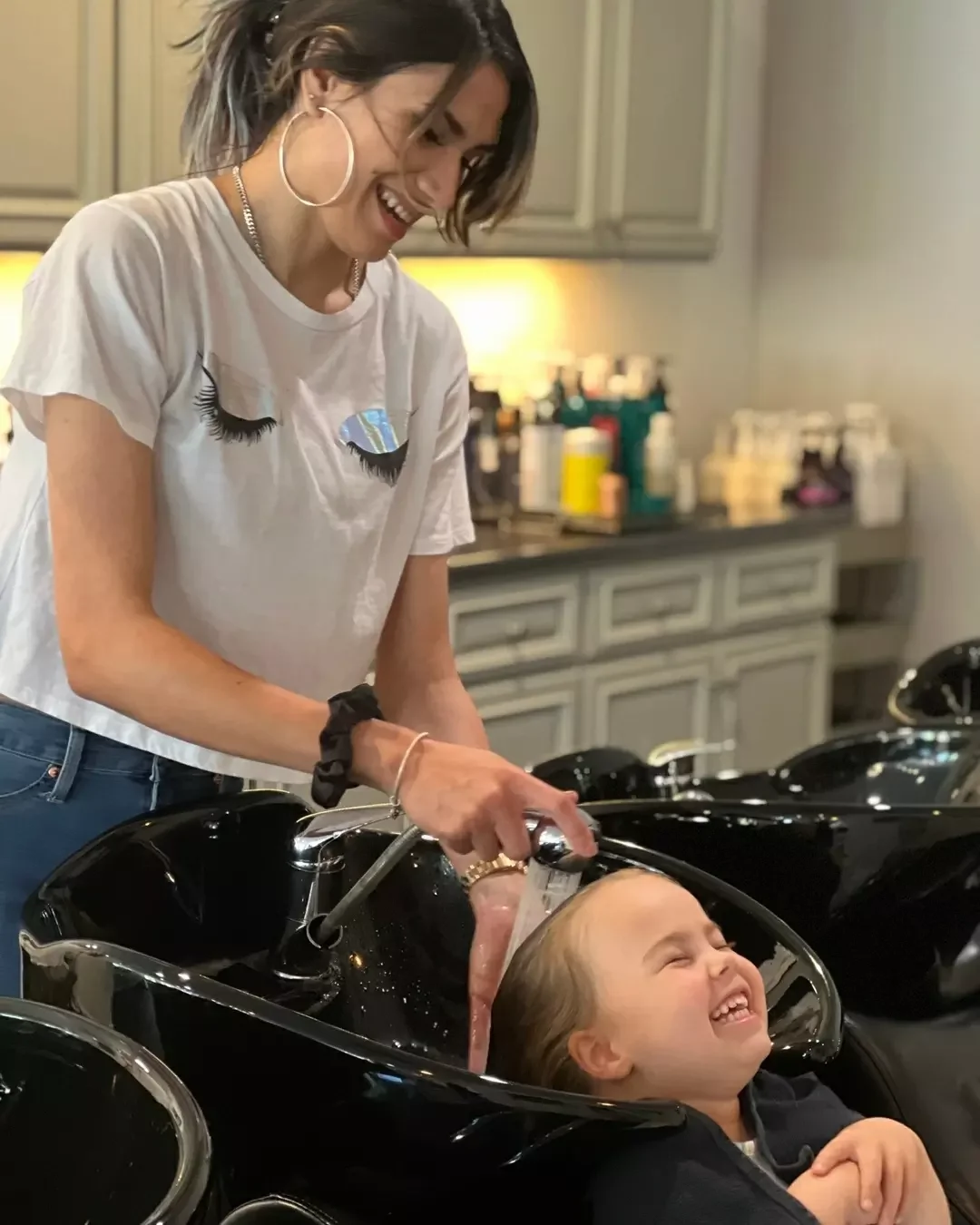 A woman washing a child's hair at a salon sink, both smiling and laughing.