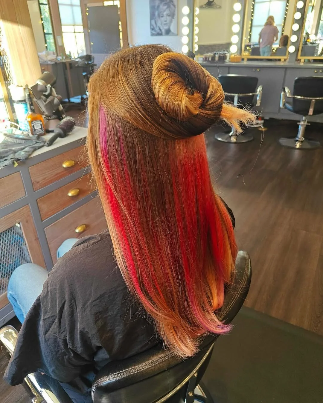 Back view of a woman in a salon chair with long, straight hair colored in shades of red, orange, and pink, styled with a vintage 1940s roll at the crown.