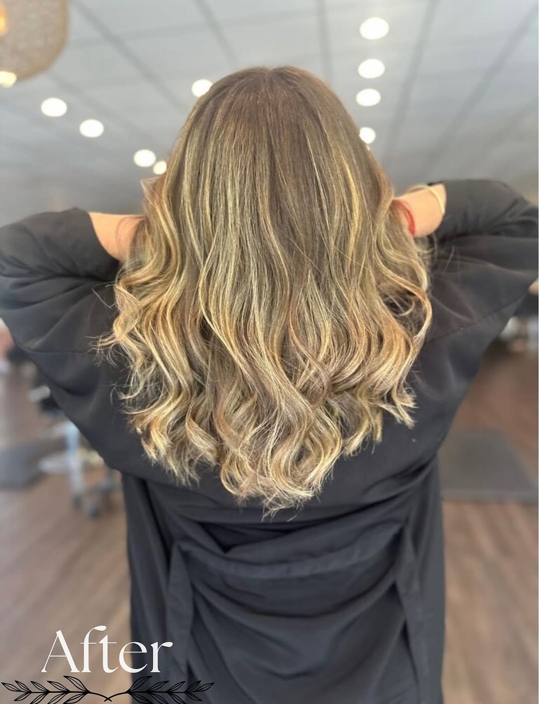 Back view of a woman with shoulder-length wavy hair featuring blended blonde highlights and light brown base color, styled in loose waves, in a salon setting.