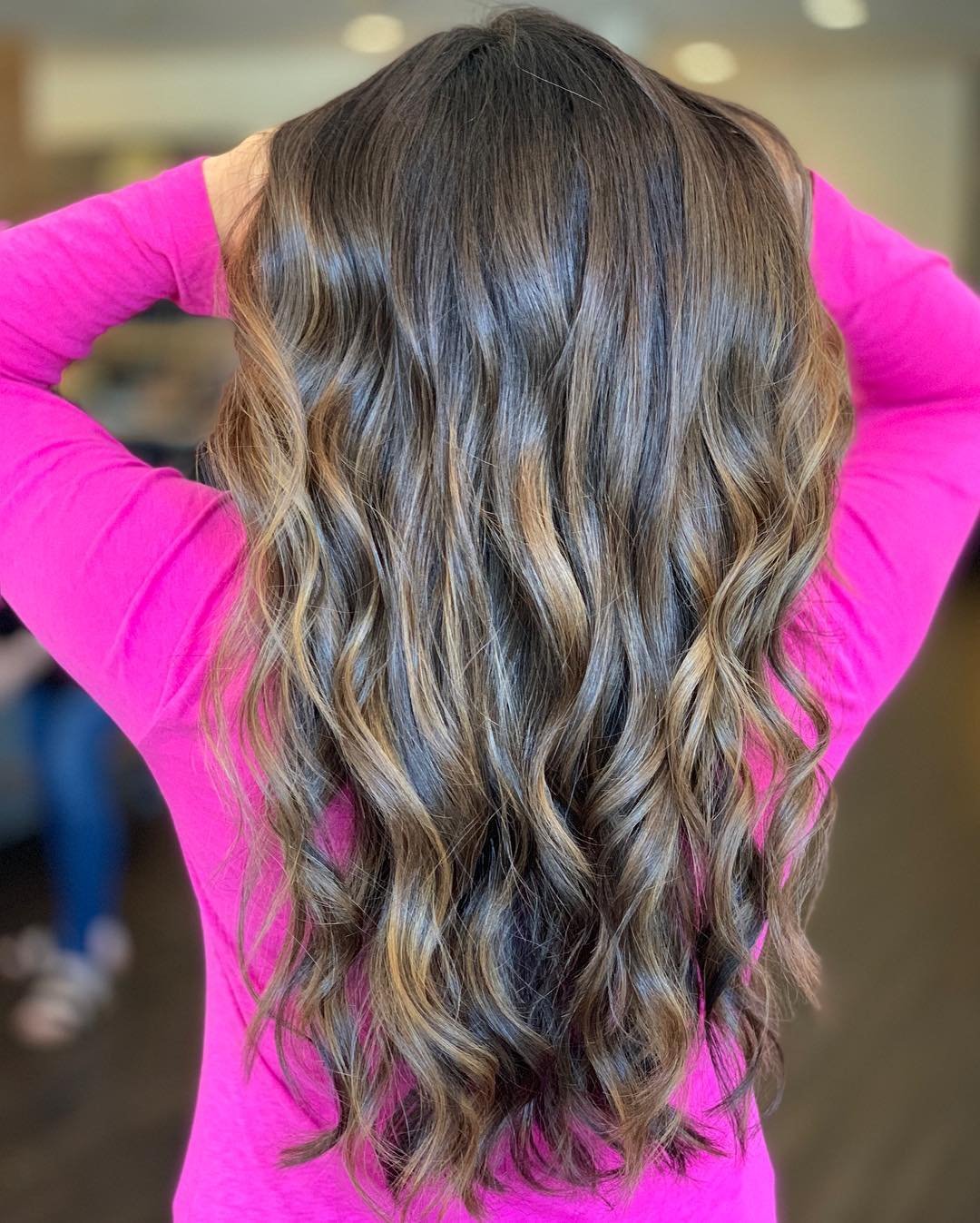 Back view of a woman with long, wavy, highlighted hair wearing a bright pink top. She has her hands raised behind her head.