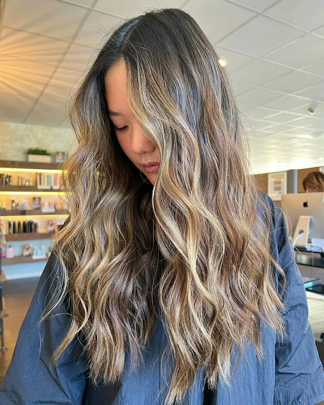 A woman with long wavy hair featuring blonde highlights and brown lowlights, gazing downward in an indoor setting with shelves and books in the background.