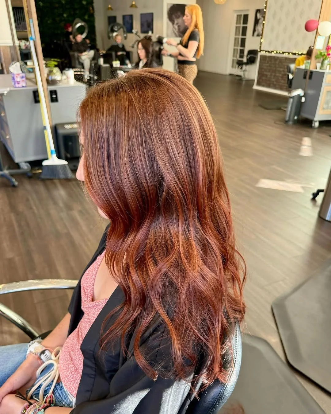 Woman with wavy, reddish-brown hair sitting in a salon chair with clients and stylists in the background.