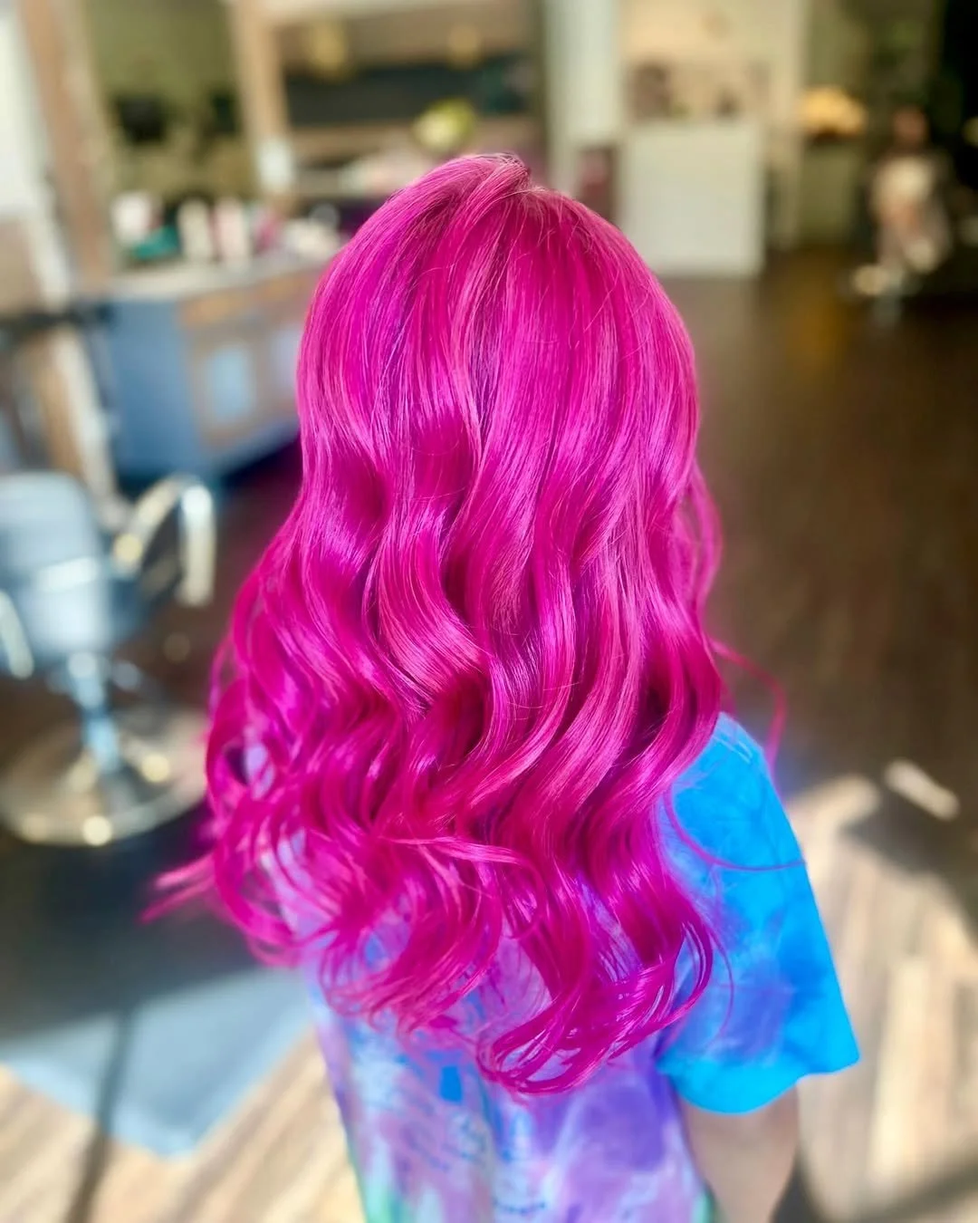 Back view of a person with bright pink, wavy hair, wearing a colorful shirt, inside a hair salon.