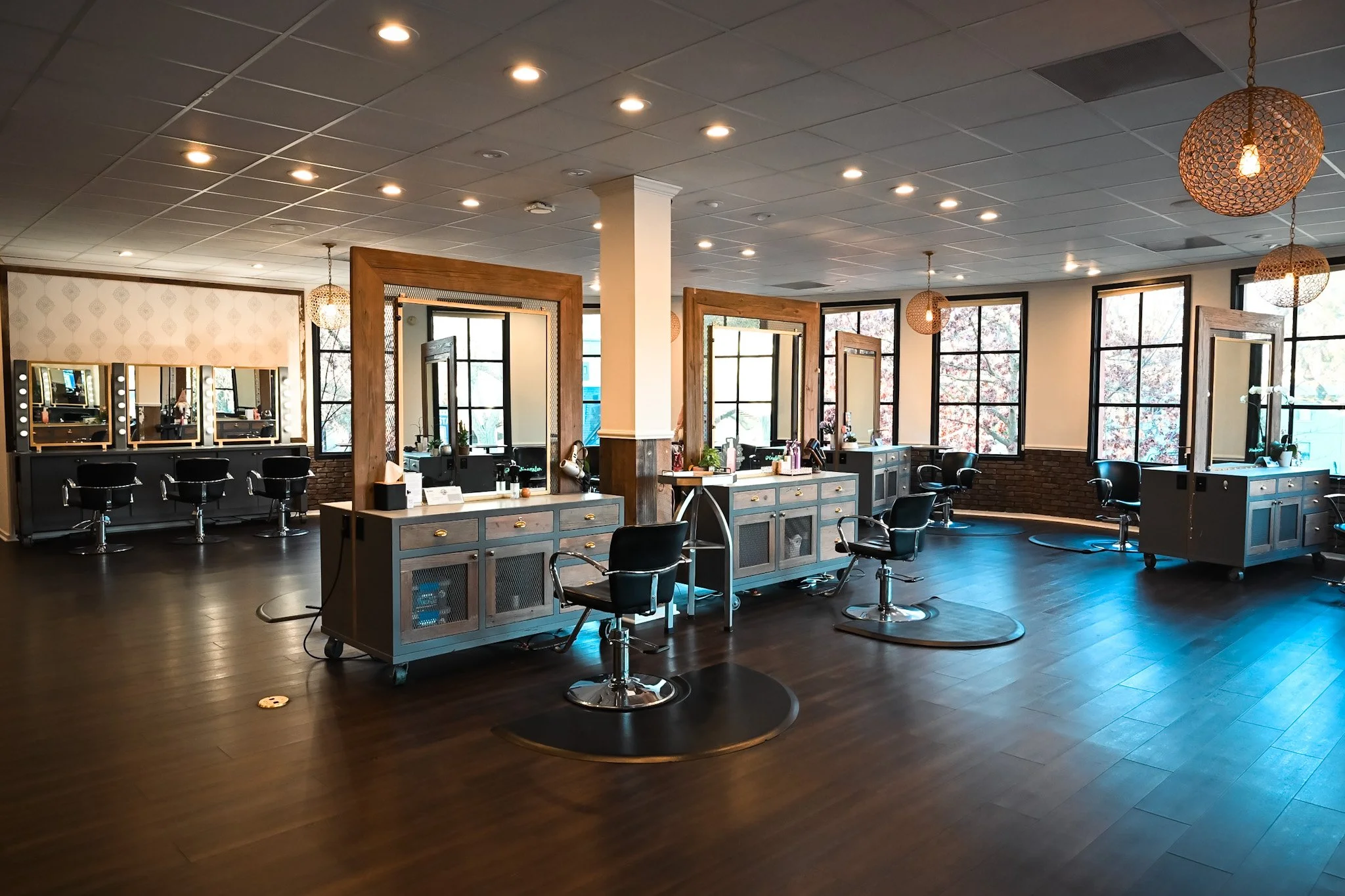 A hair salon with multiple styling stations, large windows, and ceiling lights. There are mirrors with lights, chairs, and hair styling products on the counters. The space has a modern, welcoming atmosphere with wood accents and dark flooring.