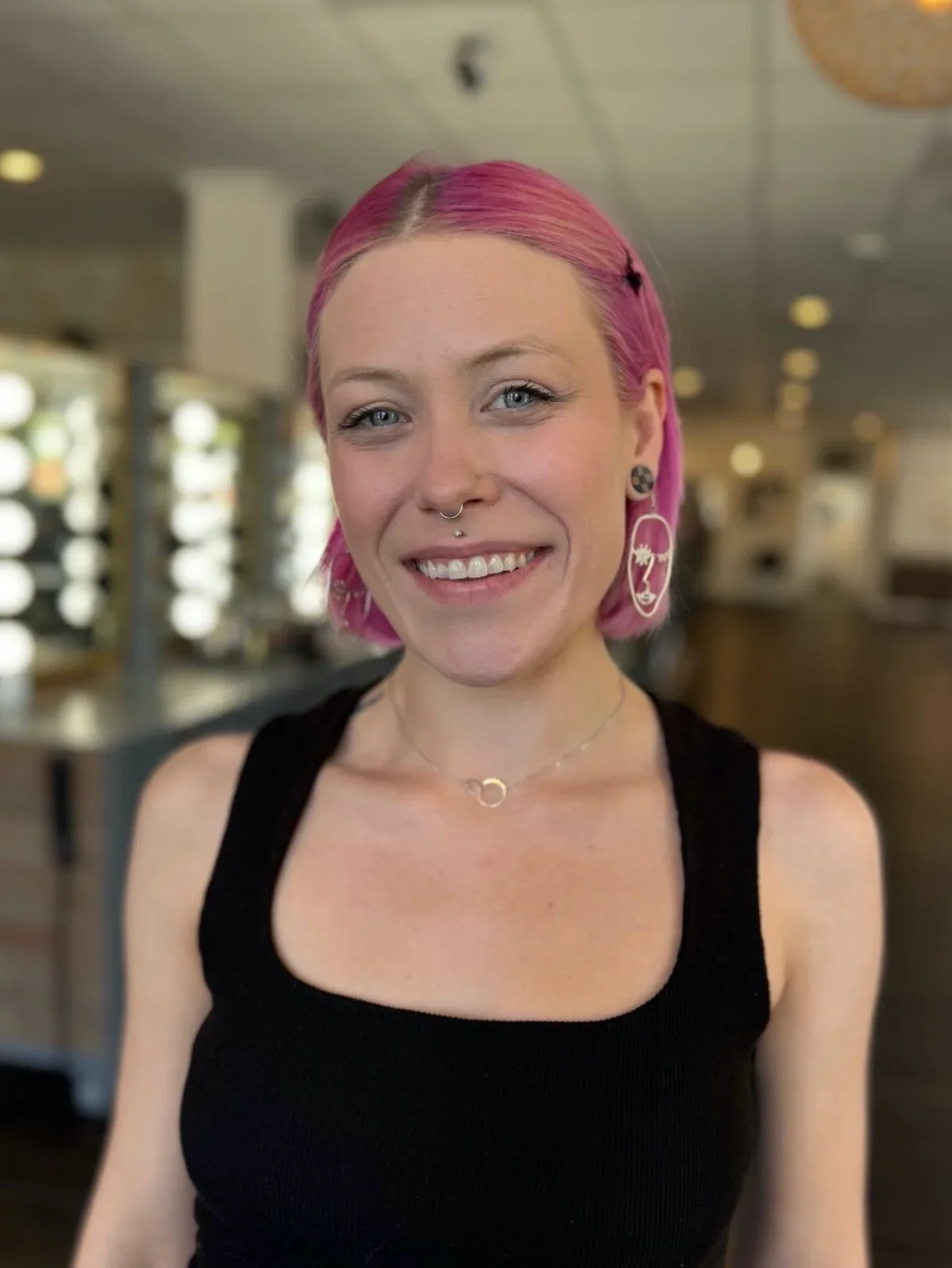 A woman with pink hair, light skin, and a friendly smile, wearing a black sleeveless top, earrings, and a necklace, standing indoors with a blurred background.