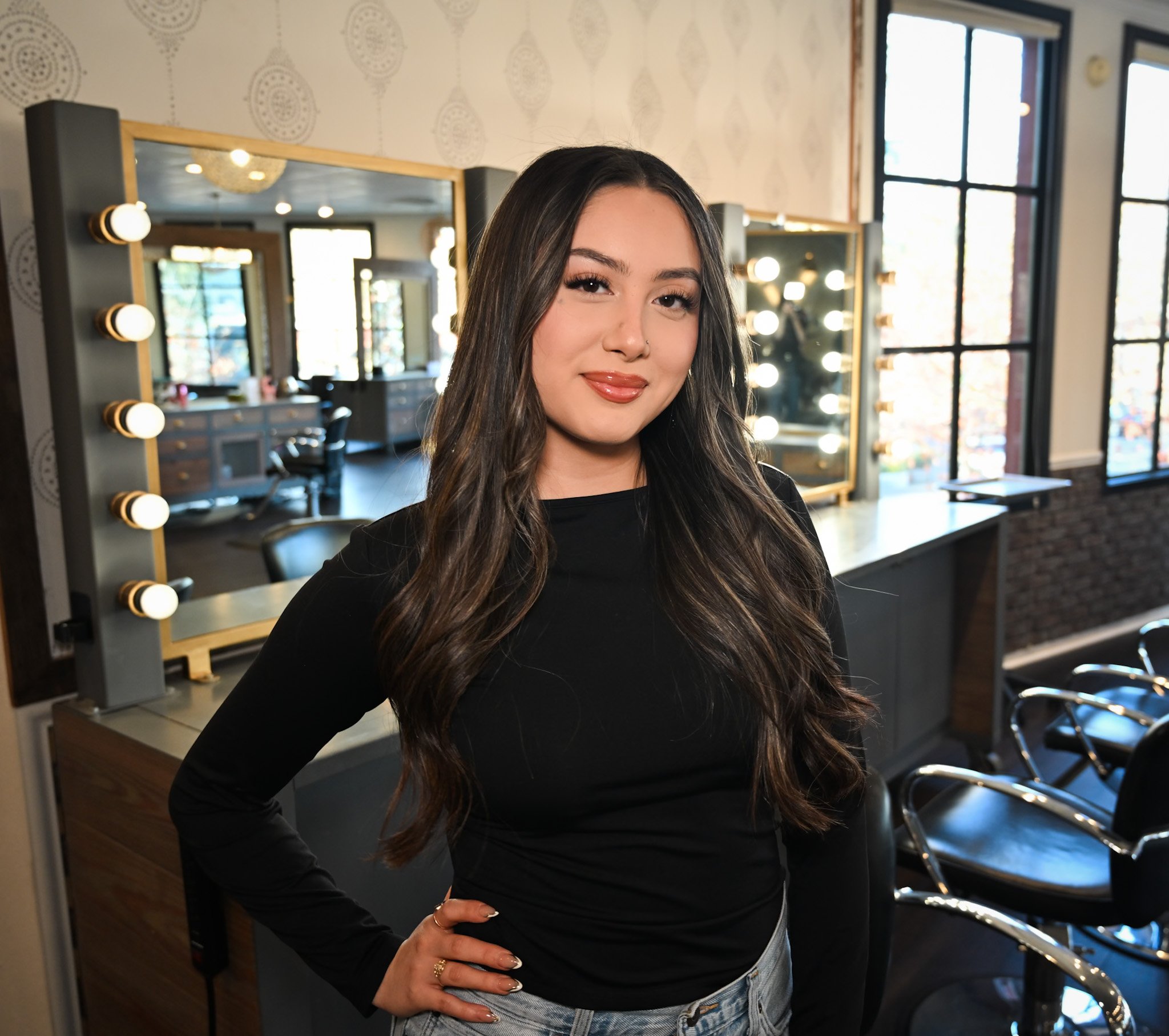 A young woman with long dark hair and a nose piercing, wearing a black shirt and jeans, poses with her hand on her hip in front of a mirror with lights, inside a modern room with large windows.