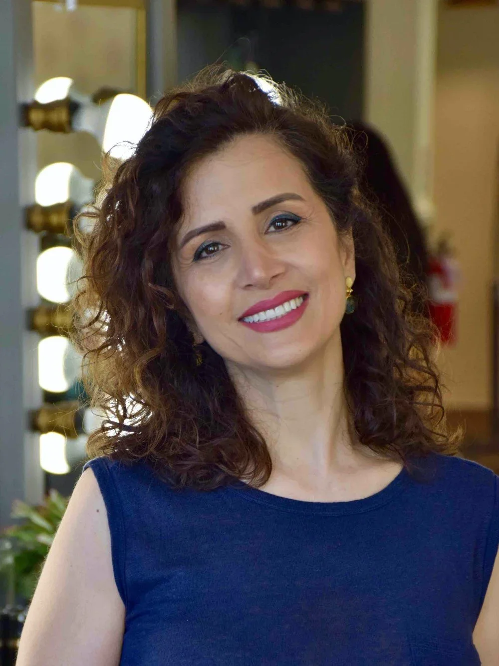 Woman with curly brown hair smiling, wearing a sleeveless navy blue top, indoors with bright lights and blurred background.