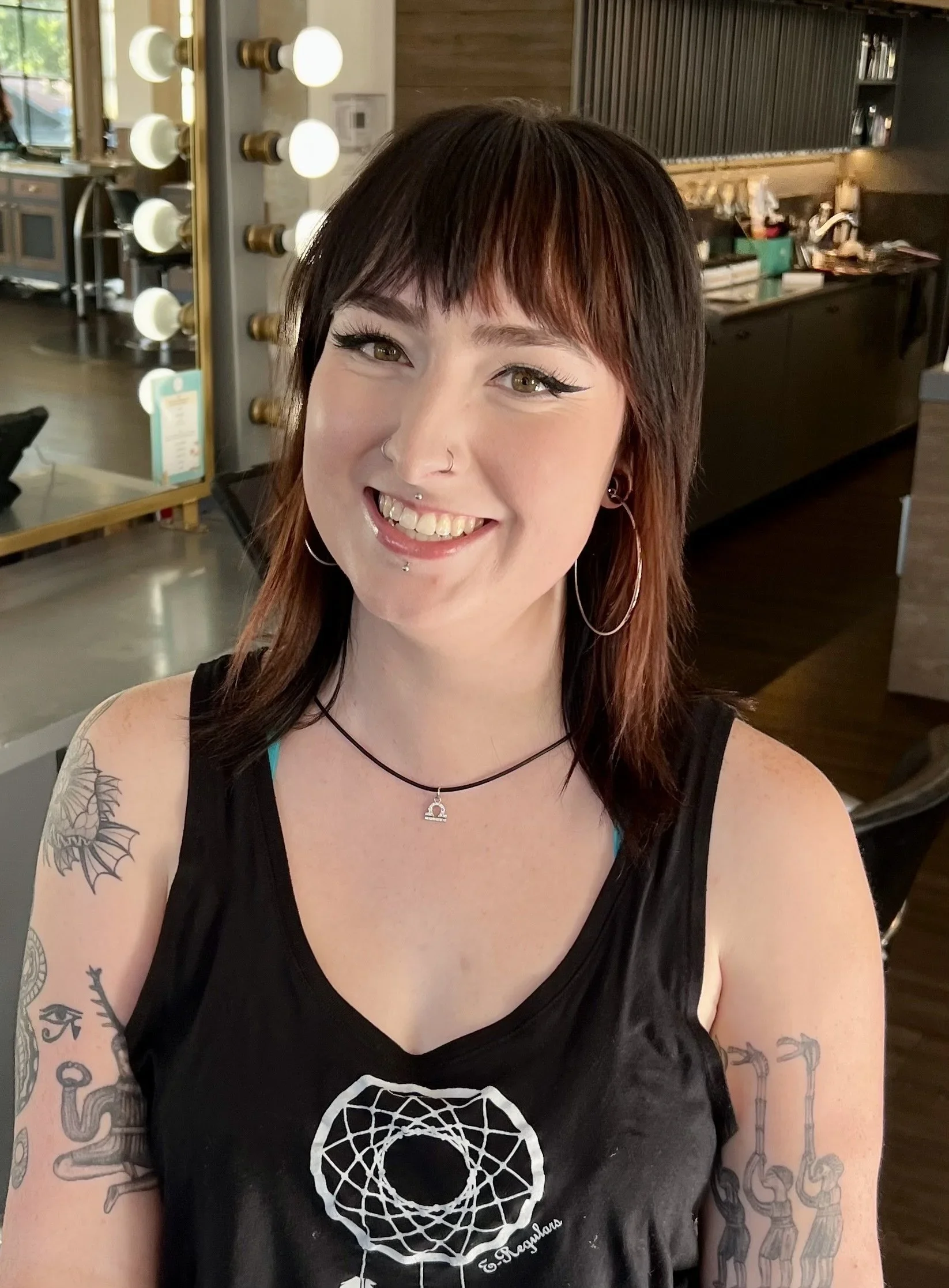 A young woman with dark brown hair with reddish highlights, smiling, in a black sleeveless shirt with a white geometric design on the front, tattoos on both arms, hoop earrings, a nose ring, and a necklace, inside a modern cafe or restaurant with a mirror, lights, and a wooden counter in the background.