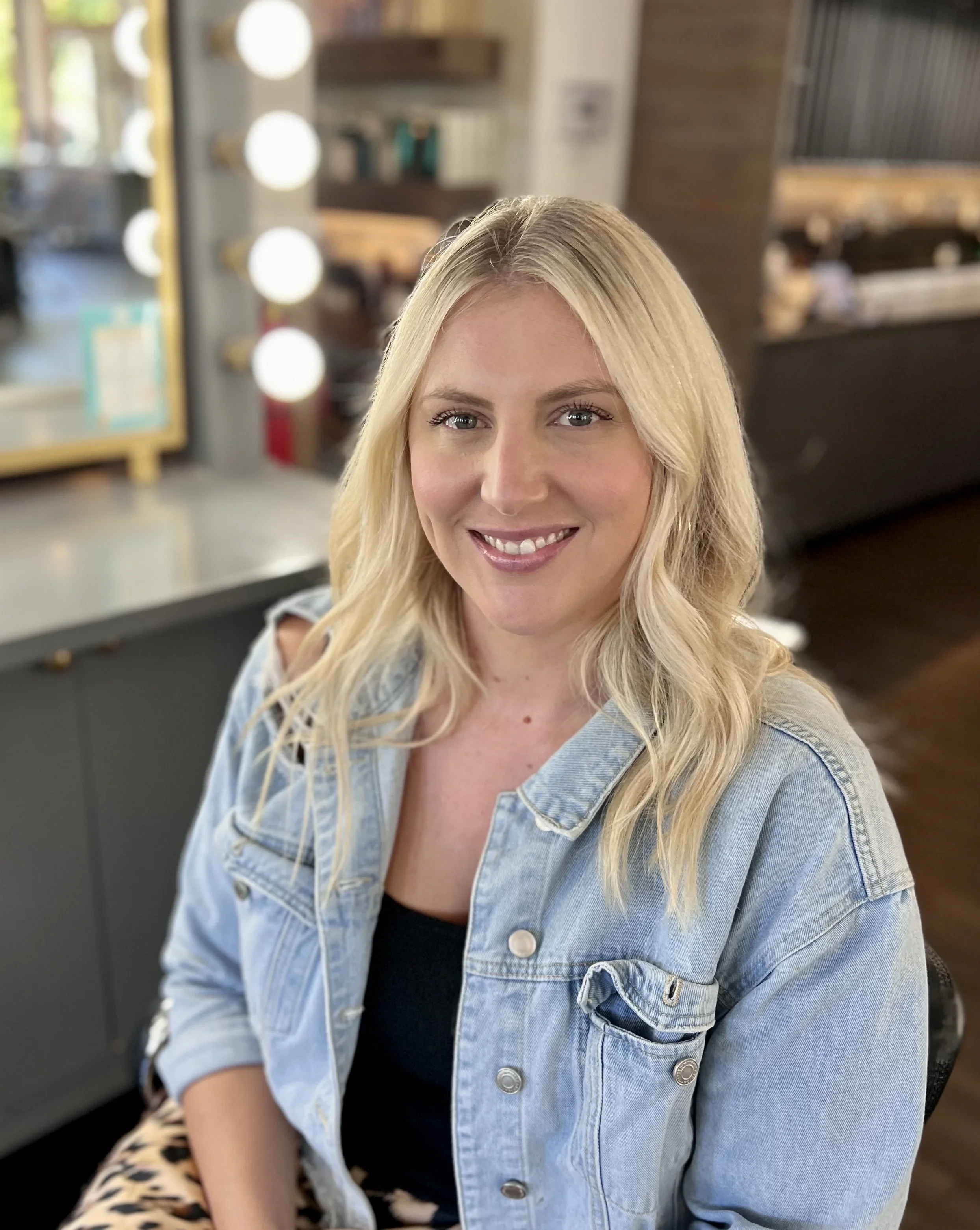 A woman with blonde hair, smiling, wearing a light denim jacket and black top, sitting in a room with a makeup mirror and warm lighting.