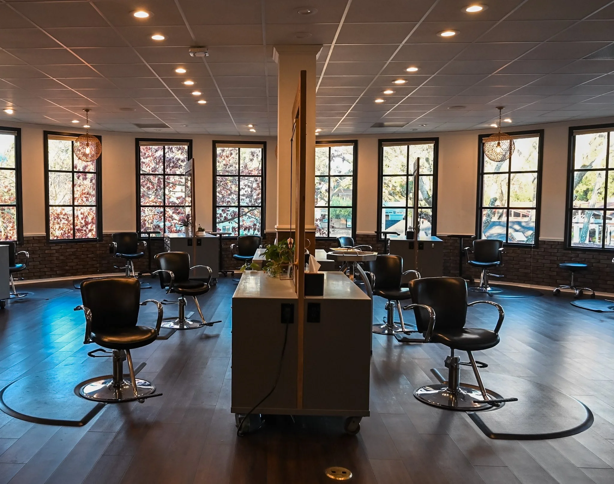 An empty haircut or beauty salon with black chairs, large windows, hanging lamps, and a divider in the middle of the room.