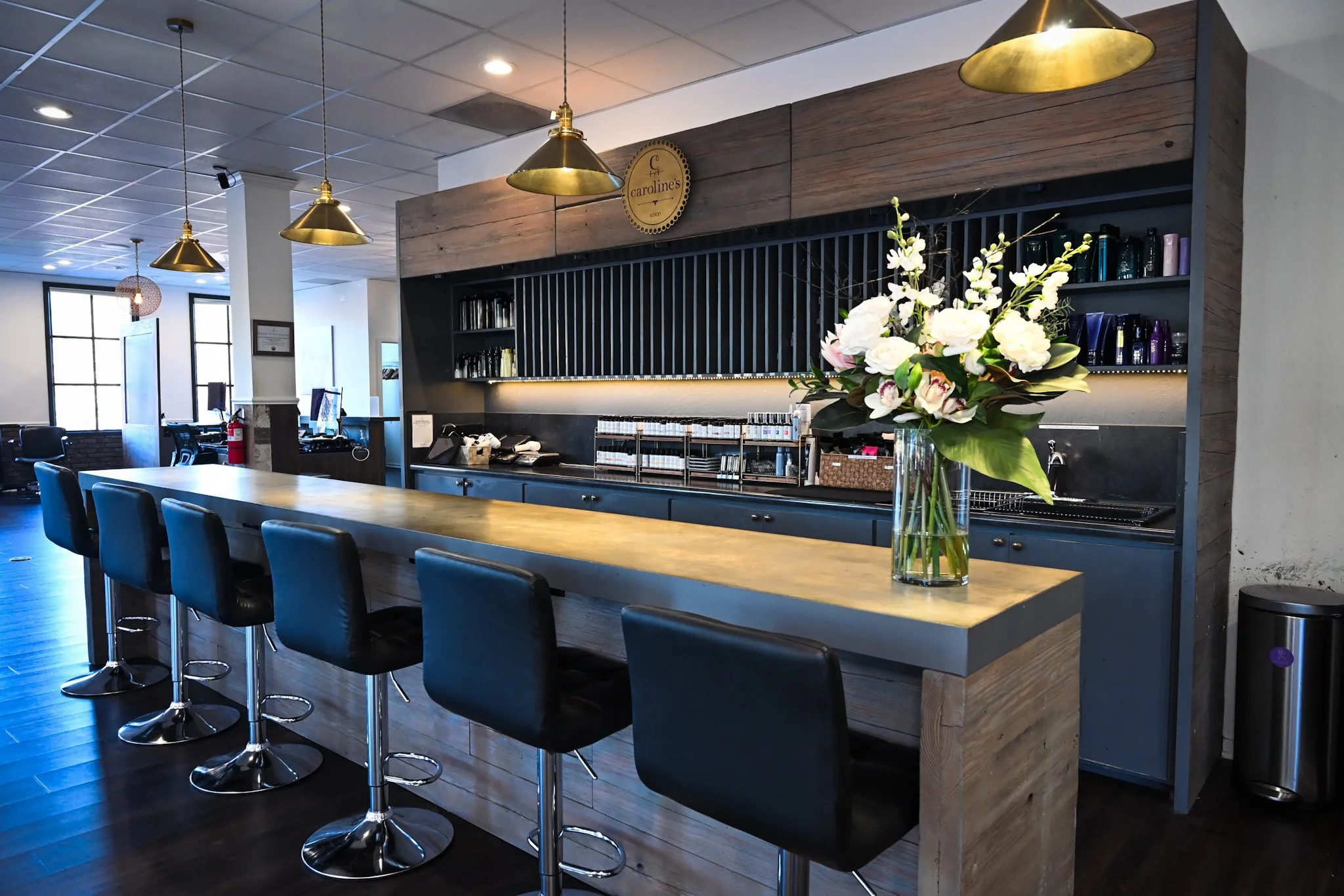 Interior of a modern salon with a long wooden counter, black high chairs, a vase with white flowers, brass pendant lights, and shelves with hair products.