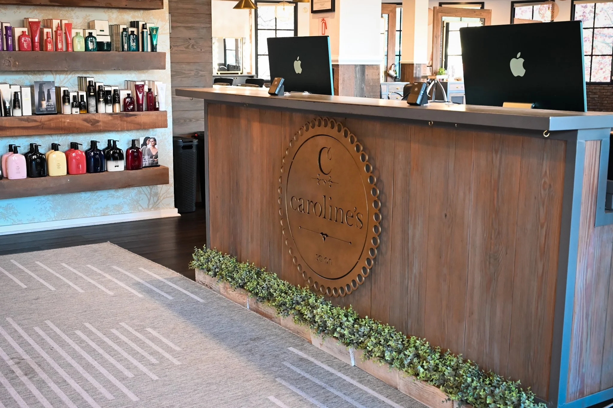 Interior of a salon reception area with a wooden desk branded 'Caroline's Salon', two Apple iMac computers, shelves with hair care products, and windows letting in natural light.