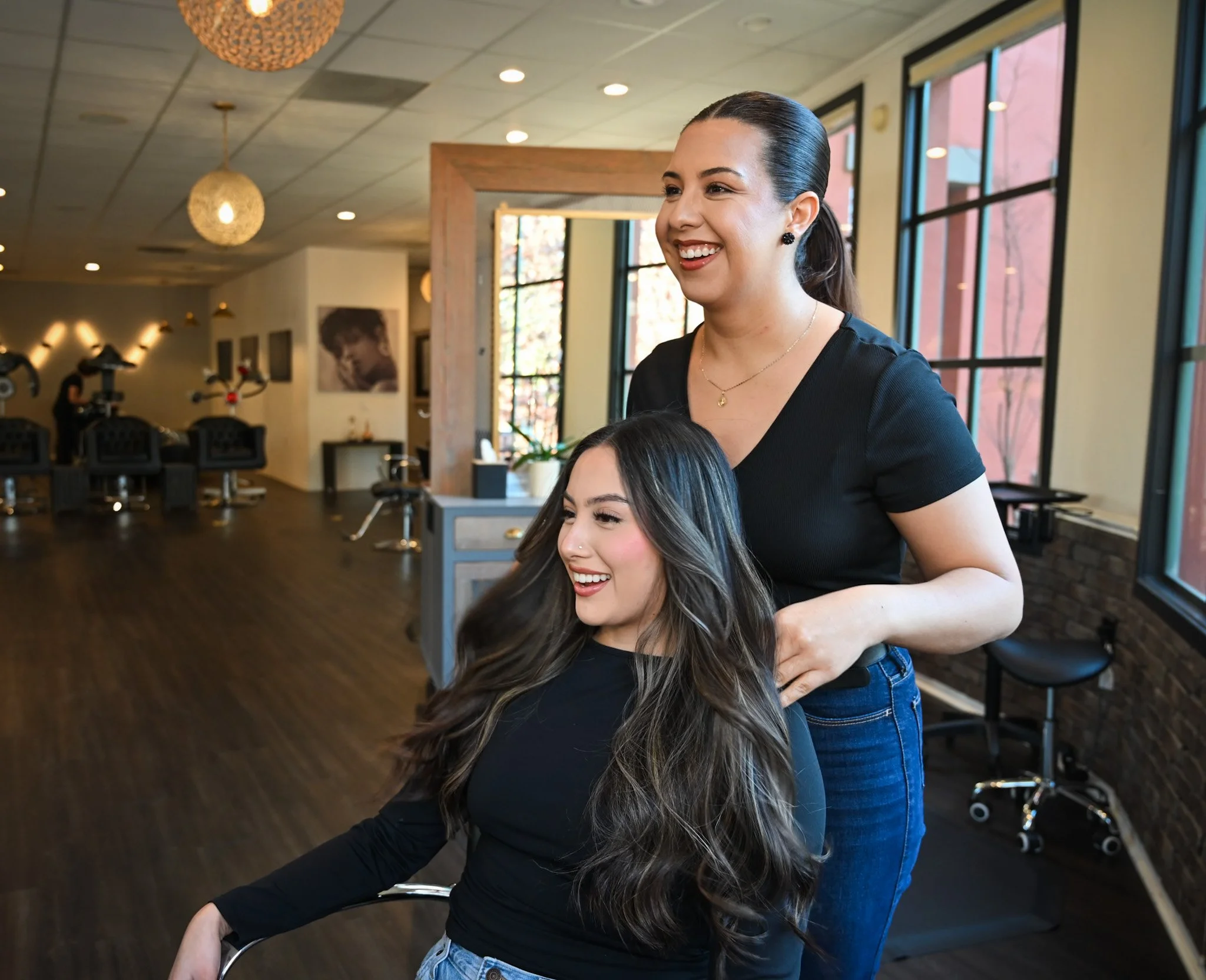 A hairstylist is smiling while working on a woman's long, wavy dark hair inside a modern salon.