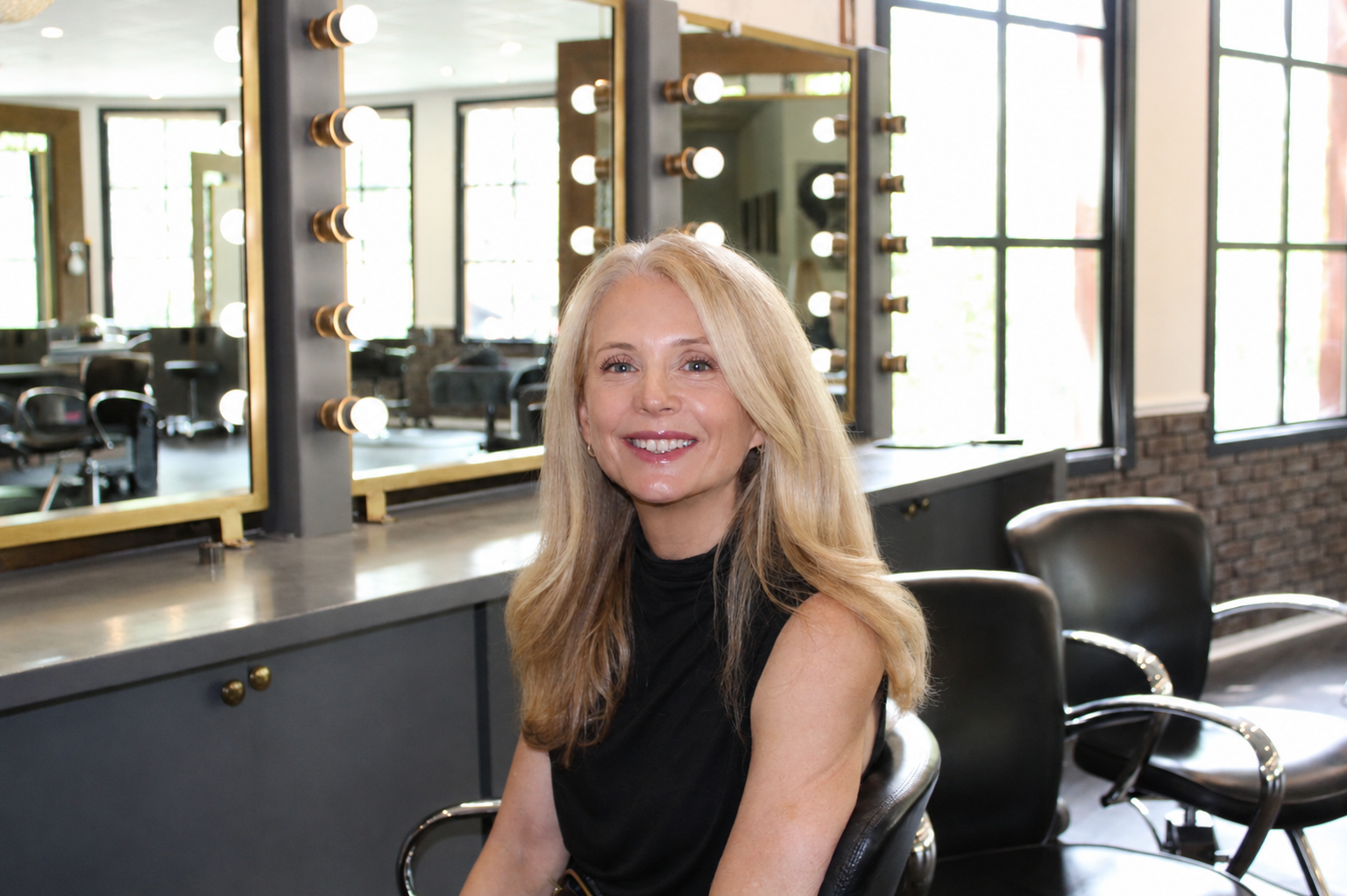 A smiling woman with long blonde hair sitting in a salon or makeup studio with large mirrors and illuminated light bulbs around them. The background features large windows with natural light and a brick wall.